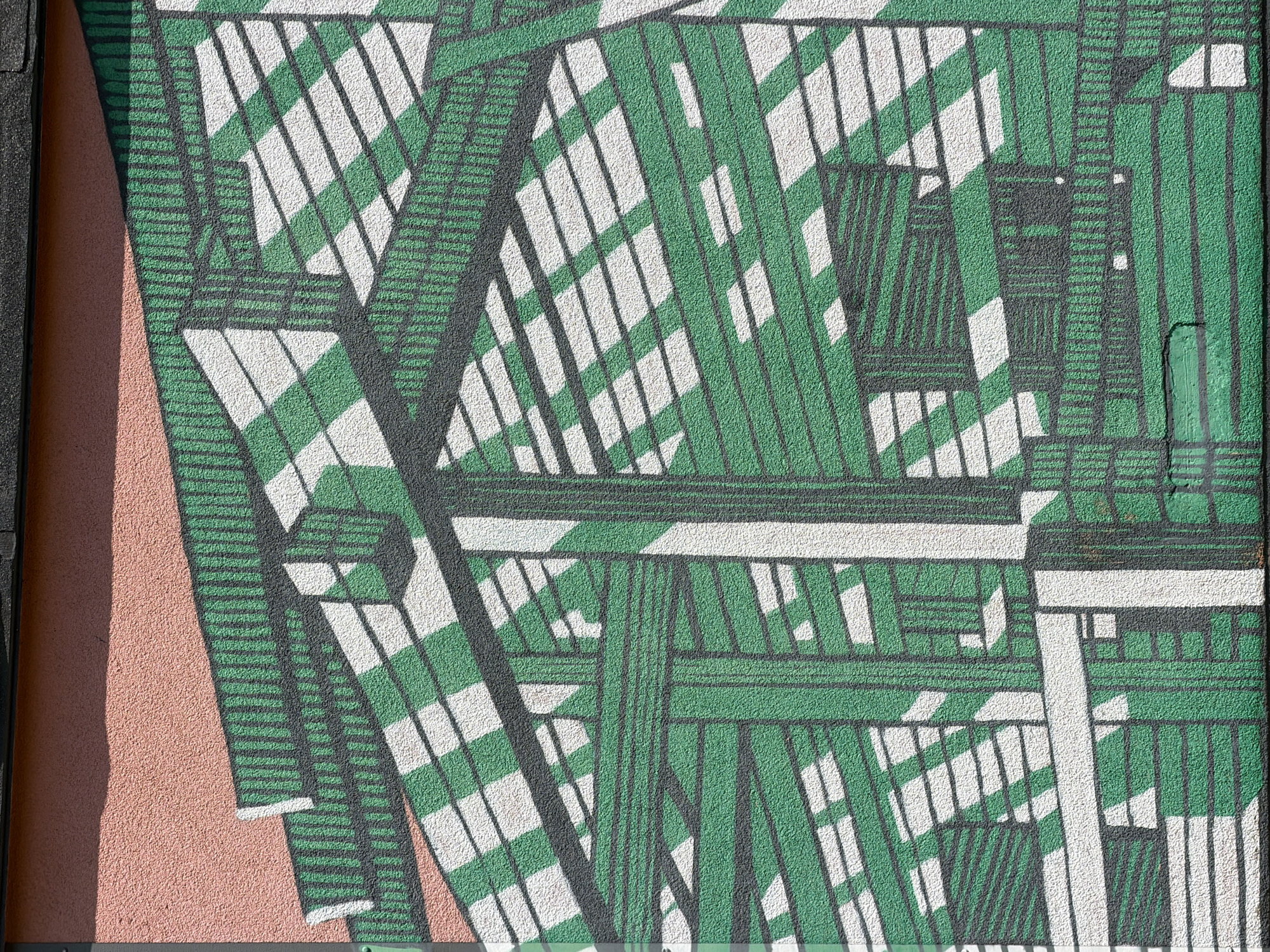 The image depicts a mural on a textured building facade, likely an external wall. The artwork features a geometric design with prominent green and white stripes, creating a sense of depth and perspective resembling a structure or architectural element, possibly an abstract representation of scaffolding or a greenhouse. Dark gray lines delineate the shapes and add to the structural illusion. The top portion of the wall has a peach-colored textured finish. A dark horizontal element, possibly a roof overhang or gutter, is visible at the very top. The overall impression is of an urban outdoor setting. The location is identified as Yekaterinburg, Russia. There are no people or discernible living subjects in the image.