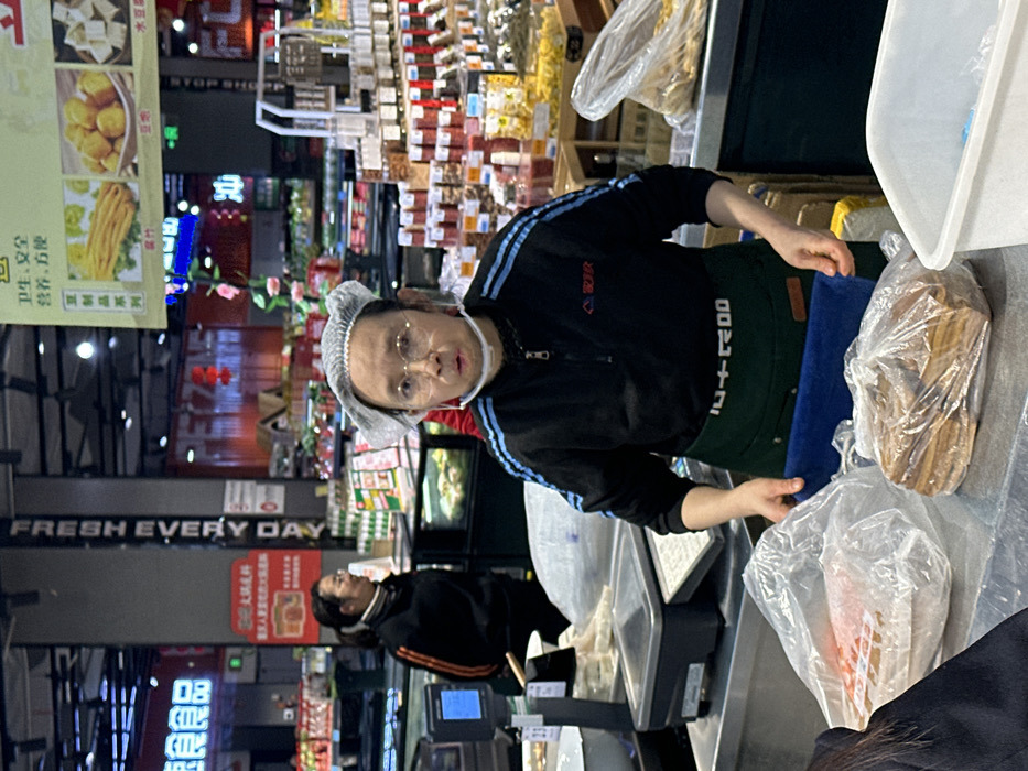 The scene depicts an interior view of a supermarket in Jiangjin, China. In the foreground, a person of East Asian descent, appearing middle-aged, is positioned centrally behind a stainless-steel counter. This individual wears round glasses, a white disposable hairnet, a black long-sleeved shirt with light blue stripes on the sleeves, and a dark green apron with white text. A clear chin strap or partial face shield is visible below their chin. Their hands are on the counter, near a blue cutting mat, attending to two clear plastic bags containing food items; one bag on the left contains lighter-colored items, and the bag on the right holds numerous stacked, rectangular, brown food items. To the left of this individual, a digital scale with a white bowl is visible. To their right, a white plastic container is present. The primary individual is looking directly towards the viewer with a slightly open mouth.
In the mid-ground, to the left and slightly behind the main subject, another East Asian female is visible, wearing a black long-sleeved top with an orange stripe on the arm and glasses. She is smiling and looking towards the left, away from the camera.
The background reveals a bustling supermarket environment with various display cases and shelves stocked with products. A prominent vertical sign on a pillar reads "FRESH EVERY DAY" in white English text. Overhead, a large hanging banner displays images of food items such as fried dough sticks, fried balls, and tofu, with Chinese text including "卫生, 安全" (Hygiene, Safety) and "豆制品系列" (Soy products series). A red section in the background features the text "PETZONE" and some red lantern-like decorations. Fluorescent lighting illuminates the entire area.
