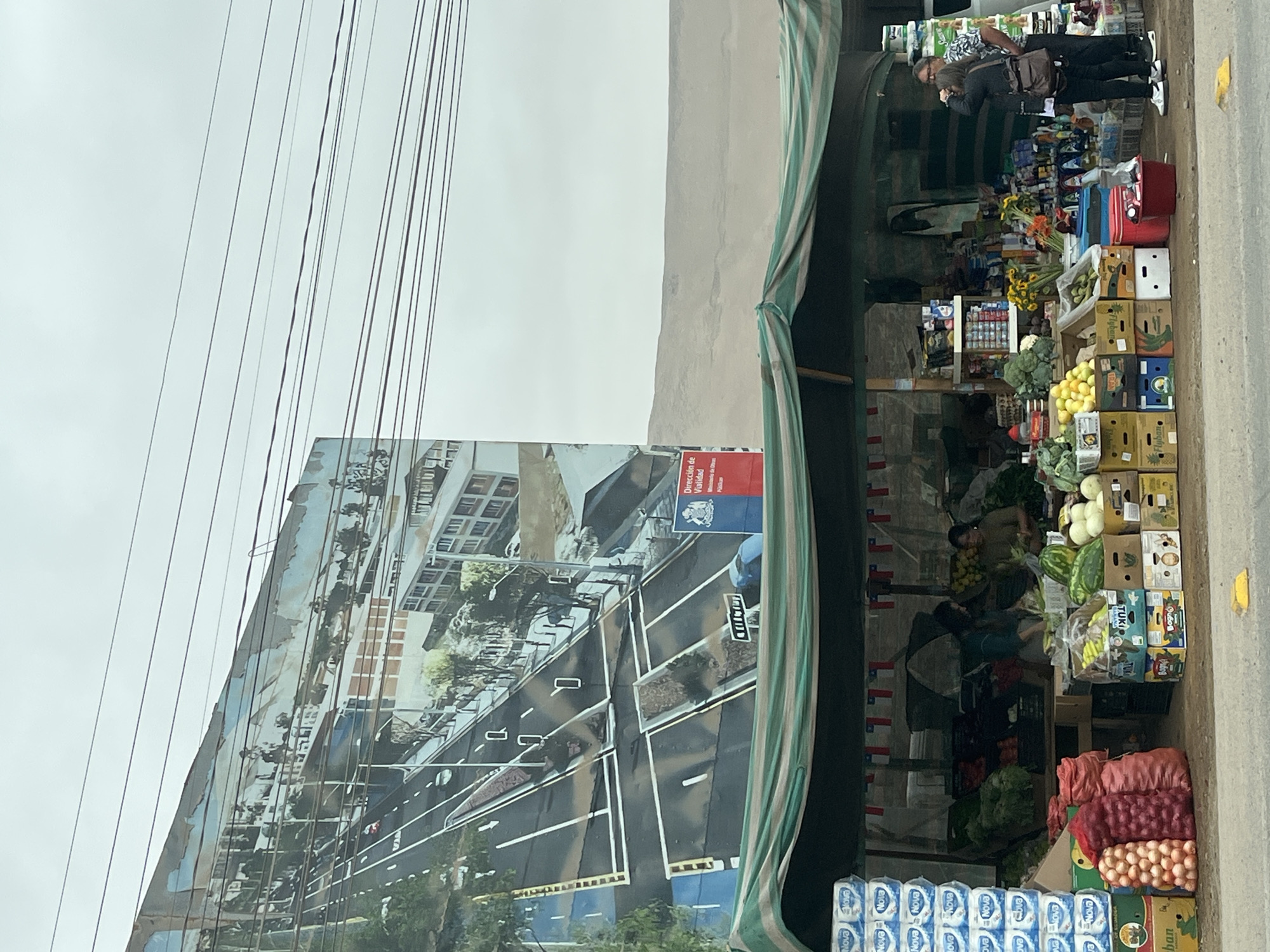 The image depicts a street scene in what appears to be Arica, Chile, showing a market stall selling produce and other goods. A person is present at the market stall, engaging with the products. There is also a large poster or billboard visible, displaying an image of the city, including buildings and roadways, with the seal "Dirección de Vialidad." Below the produce at the front of the image are stacks of toilet paper, with brands such as Nova on the packaging.