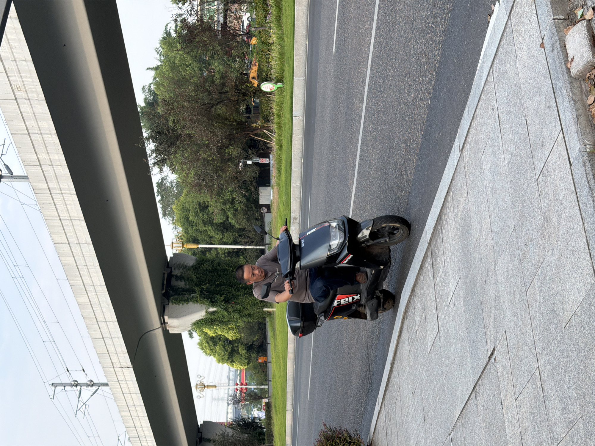 A middle-aged man wearing a gray t-shirt and blue jeans is riding a black electric scooter with a storage box on the back. The scooter has a logo on its side that reads "FEIX". He is looking forward as he rides on the right side of the road. The road is asphalt with white lane markings. To the left of the road is a sidewalk made of gray paving stones. In the background, there is a large elevated concrete structure, possibly an overpass or a bridge, with overhead power lines. Green trees and vegetation are visible beyond the road. The sky is overcast. The scene appears to be in a city environment, possibly in Jiangjin, China, given the architecture and vegetation.
