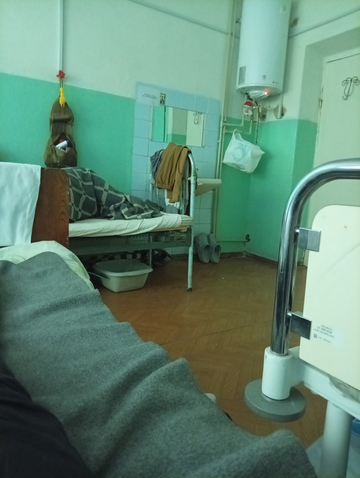 The image depicts the interior of what appears to be a hospital room, located in an Unknown, Unknown urban area. The room's color scheme features pale green walls with horizontal bands of a darker green. A hospital bed is present, partially covered with a grey blanket, and a second bed is visible in the background with a patterned blanket on top. A small hanging bag is attached to the wall beside the bed in the background. A washbasin and assorted personal items like a jacket and a pair of shoes are positioned near the bed. In the right foreground, a hospital bed frame is visible, including the foot. The floor is covered with brown tiles. A water heater hangs on the wall near the sink. The room's elements contribute to a simple and utilitarian atmosphere typical of a medical facility.