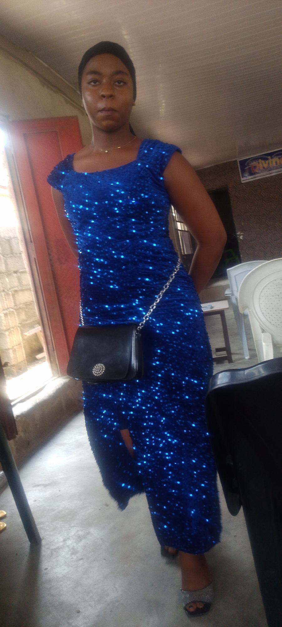 A woman stands in what appears to be an indoor space in Lokoja, Nigeria. She is wearing a blue, sequined dress with a slit. She carries a small black handbag on a silver chain slung over her hip. She wears silver sandals. A red door is visible in the background to her left, and a partial view of a white plastic chair, table, and signage that says "divine" are to her right. The surrounding area seems dimly lit with light from the window creating shadows and glare. The walls are white and dull in the background.