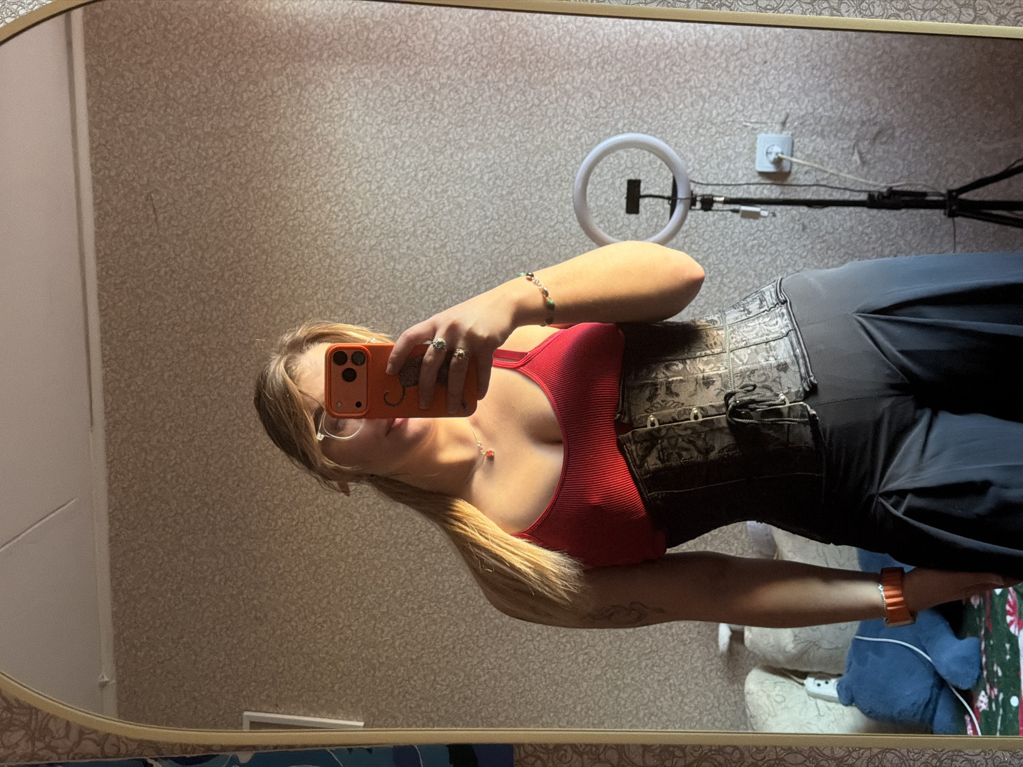 A young woman is taking a mirror selfie. She's wearing a red top under a patterned corset and black trousers. She holds an orange smartphone, obscuring her face. The background shows a patterned wallpaper, a ring light on a stand, and part of a room with some items on the floor, including a blue stuffed animal and a Christmas-themed mat. No information suggests this image was taken in Naberezhnye Chelny, Russia.