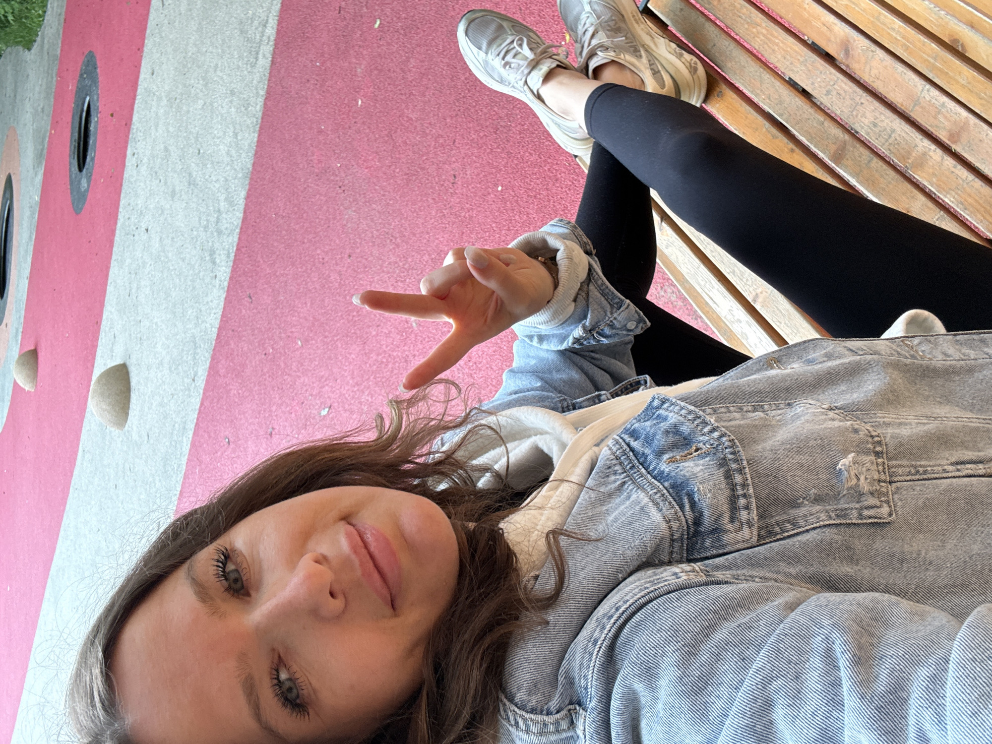 A medium shot captures a young woman with brown hair, her face turned toward the camera. She is wearing a light-wash denim jacket over a hooded sweatshirt, black leggings, and light-colored athletic shoes. Her right hand is raised, forming a peace sign. She is seated on a wooden bench. The background consists of a brightly colored, possibly artificial, surface with pink and light gray stripes, punctuated by circular openings and dome-shaped elements. The scene appears to be an outdoor recreational area, possibly a playground or park, though a specific location in Moscow, Russia, cannot be identified from the visual information provided.