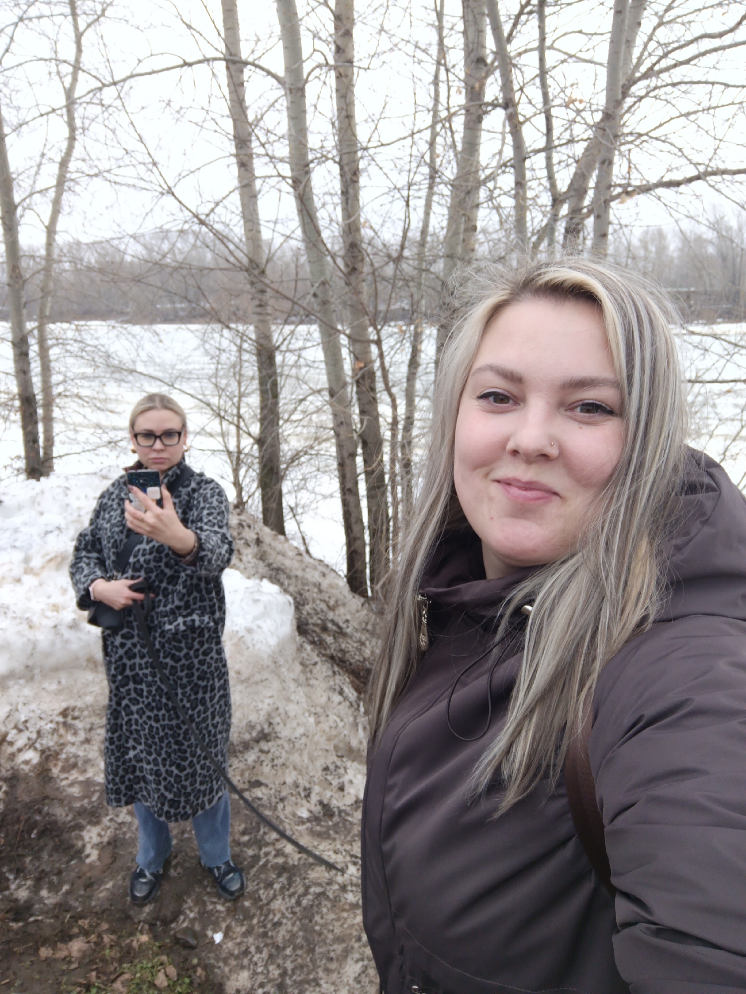Two women are outdoors in a winter environment. One woman is closer to the camera, wearing a brown hooded jacket with blonde hair streaked with lighter highlights. She has a visible nose piercing. The second woman is further back, wearing a long gray and black leopard-print coat, glasses, and jeans, holding a smartphone. The ground is a mix of mud and snow, and there is a large snow pile. The background features bare trees and a landscape of snow and water. The location is Chaikovsky, Russia.