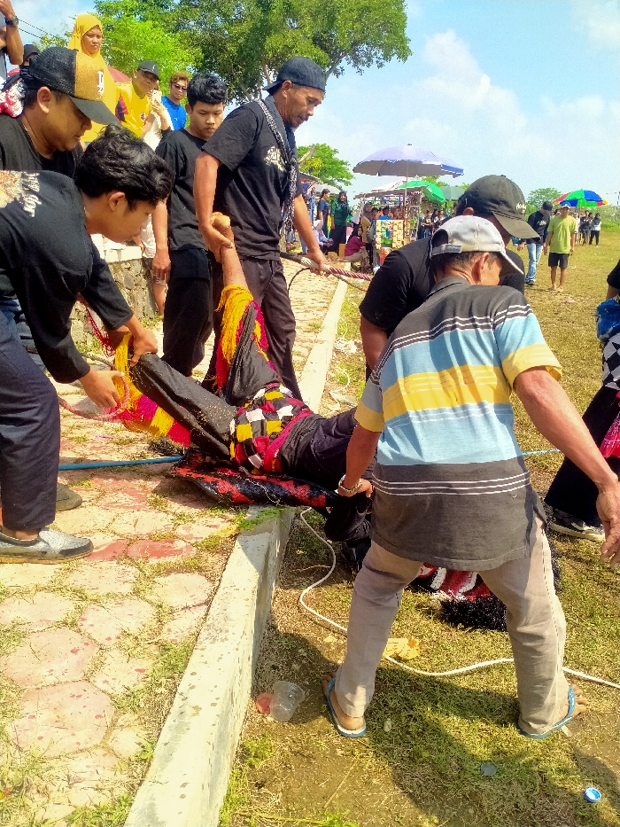 An outdoor scene in Celep, Indonesia, depicts multiple individuals assisting a person lying prone on the ground near a concrete curb. The person on the ground wears dark pants and a patterned top with red, yellow, black, and white squares/strips, with additional yellow and red fabric strips around their limbs. They are resting on a dark, possibly wet, mat or cloth.
Six men are actively engaged with the person on the ground. On the left, a man in a black long-sleeved shirt and dark pants holds one arm of the person. Beside him, a man in a black t-shirt and dark baseball cap also grips the same arm. A man in a black t-shirt and black baseball cap, positioned on the right, holds a thick, braided rope that extends towards the prone individual. Another man in a black t-shirt and black baseball cap is visible directly behind him. In the immediate right foreground, an older man in a striped blue, yellow, and dark grey t-shirt, light brown pants, and flip-flops, bends over, holding one of the prone person's legs. A seventh man in a yellow t-shirt and dark cap is visible in the background, facing the group.
The ground consists of reddish-brown hexagonal pavers on the left, separated by a concrete curb from an area of dry grass and dirt on the right. A clear plastic bottle, a blue plastic cap, and another yellow plastic object are discarded on the grass. In the mid-ground, a crowd of people gathers around several market stalls topped with colorful umbrellas (blue, green, white, and red). Green foliage from trees is visible in the upper background against a clear blue sky with sparse white clouds.