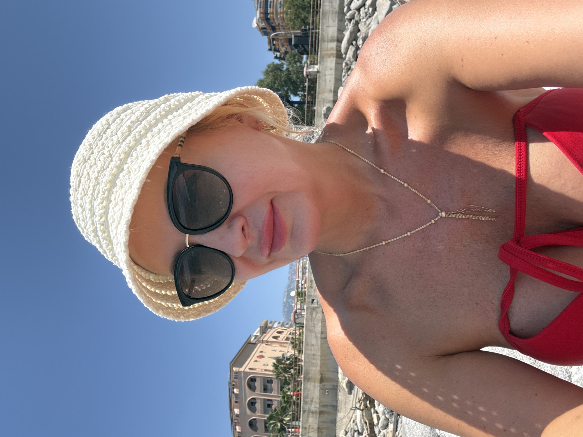 A close-up, selfie-style shot captures a blonde woman wearing a white crocheted bucket hat and dark sunglasses. She has on a red bikini top and a gold necklace with a tassel pendant. The woman is positioned in the foreground, with the background showing a clear blue sky, a stone-covered beach, a concrete wall, buildings, and palm trees, suggesting a Mediterranean or coastal location. The image appears to be taken outdoors on a sunny day. The buildings in the background are Mediterranean in style, with arched windows and balconies. One building has a sign with text that is partially visible.
