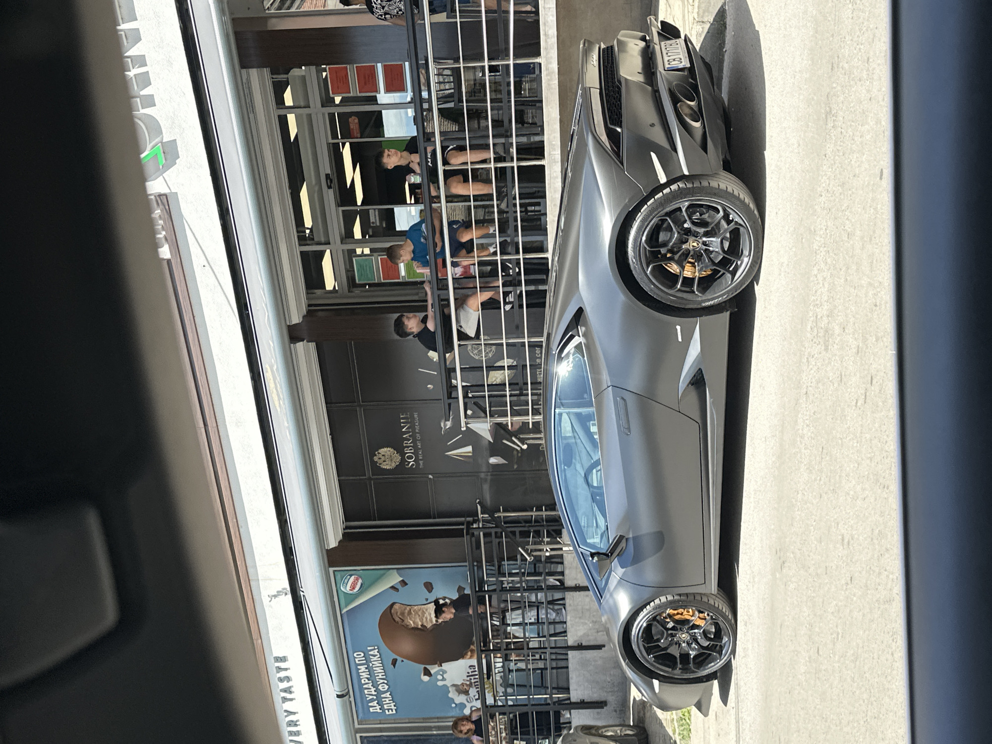 The image captures a street scene in Veliko Tarnovo, Bulgaria, with a matte gray Lamborghini Huracan parked on the asphalt. In the background, a building features an outdoor seating area with several people. A large advertisement poster is visible on the left side of the building, depicting a broken chocolate ice cream bar with the text "ДА УДАРИМ ПО ЕДНА ФУНИЙКА!" which translates to "Let's go for a cone!" and "familia" in smaller text. To the right of the poster, the entrance to a business called "SOBRANJE" is visible, with the tagline "THE REAL ART OF PLEASURE" below it. Several young men are seated at tables in the outdoor seating area, appearing to be engaged in conversation. The lighting suggests it is daytime, with sunlight casting shadows. The overall scene depicts a vibrant urban setting with a luxury sports car as a prominent element.