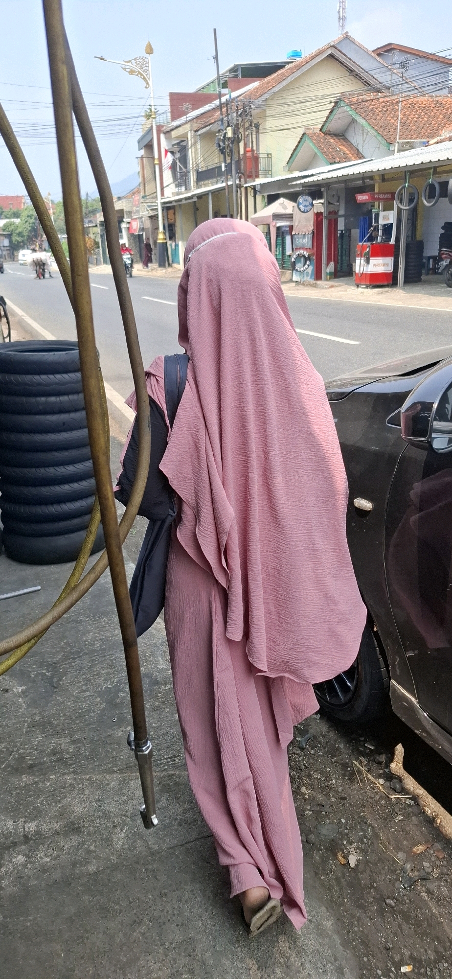 In Sukasari, Indonesia, a person wearing a pink hijab and matching abaya stands on a sidewalk next to a dark-colored car. The person is facing away from the camera, with their back towards a street that is lined with buildings and a gas station. Several stacked tires are visible on the sidewalk to the left of the person, and a metal pole with attached hoses stands in the foreground. The street appears to be a busy thoroughfare, with a few vehicles and pedestrians in the distance. The environment suggests a commercial area with a mix of residential and retail establishments.