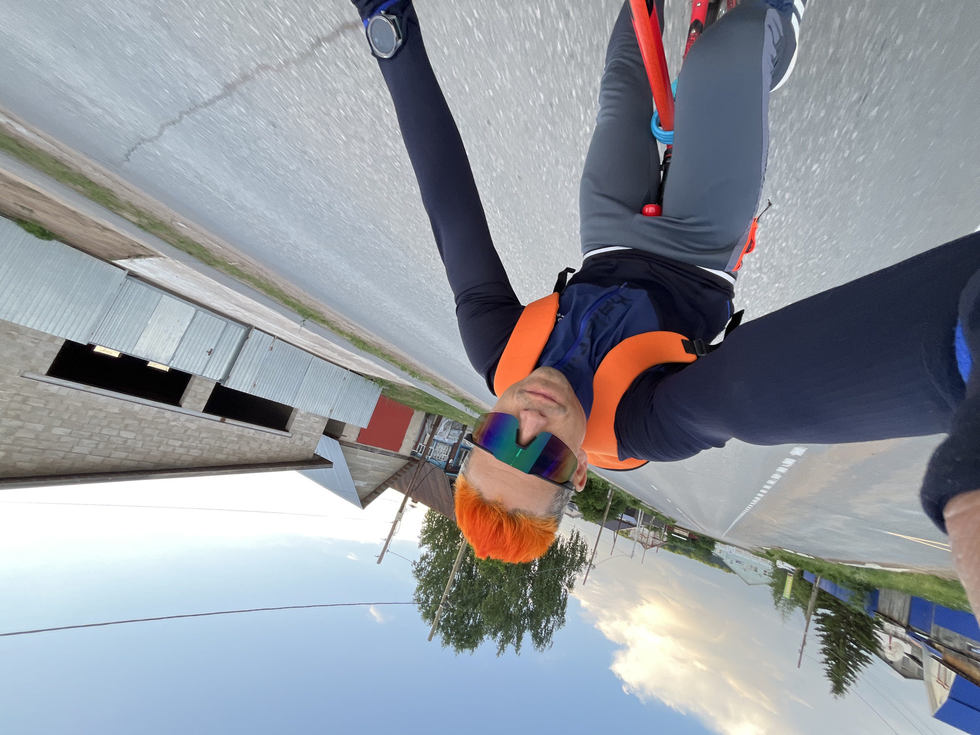 A cyclist takes a selfie while riding on a road in what appears to be a rural area. The cyclist has dyed orange hair and wears rainbow-colored sunglasses, a dark long-sleeved shirt, and an orange vest. The bicycle is red, with gray pants and white sneakers. The image orientation is upside down. The background includes a gray brick building with dark openings, a red house with a gray roof, a road with a painted white line, trees, houses, and a partly cloudy sky. This location can be recognized as a road near the city of Chaikovsky, Russia.