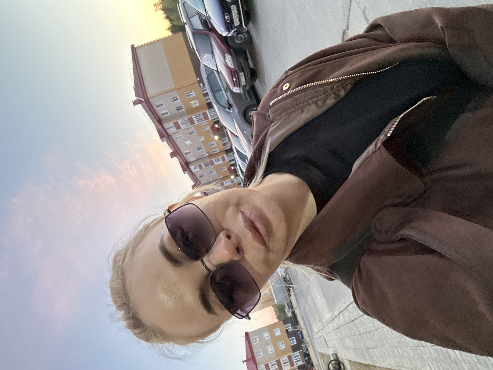 A woman with fair skin and blond hair, wearing sunglasses and a brown jacket over a black top, is taking a selfie outdoors. Behind her, there is a multi-story residential building with a white and orange facade, parked cars, and a sky with light colors. The location is Vysokiy, Russia, as inferred from the surrounding environment.