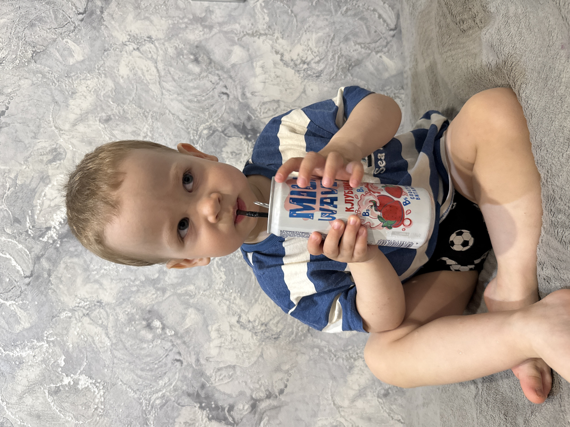 A young boy sits on a gray patterned surface, presumably a bed or sofa, drinking from a white carton with a straw. He wears a blue and white striped shirt and black shorts with soccer ball patterns. The background consists of a gray and white patterned wall. The specific location is Rusanovo, Russia.