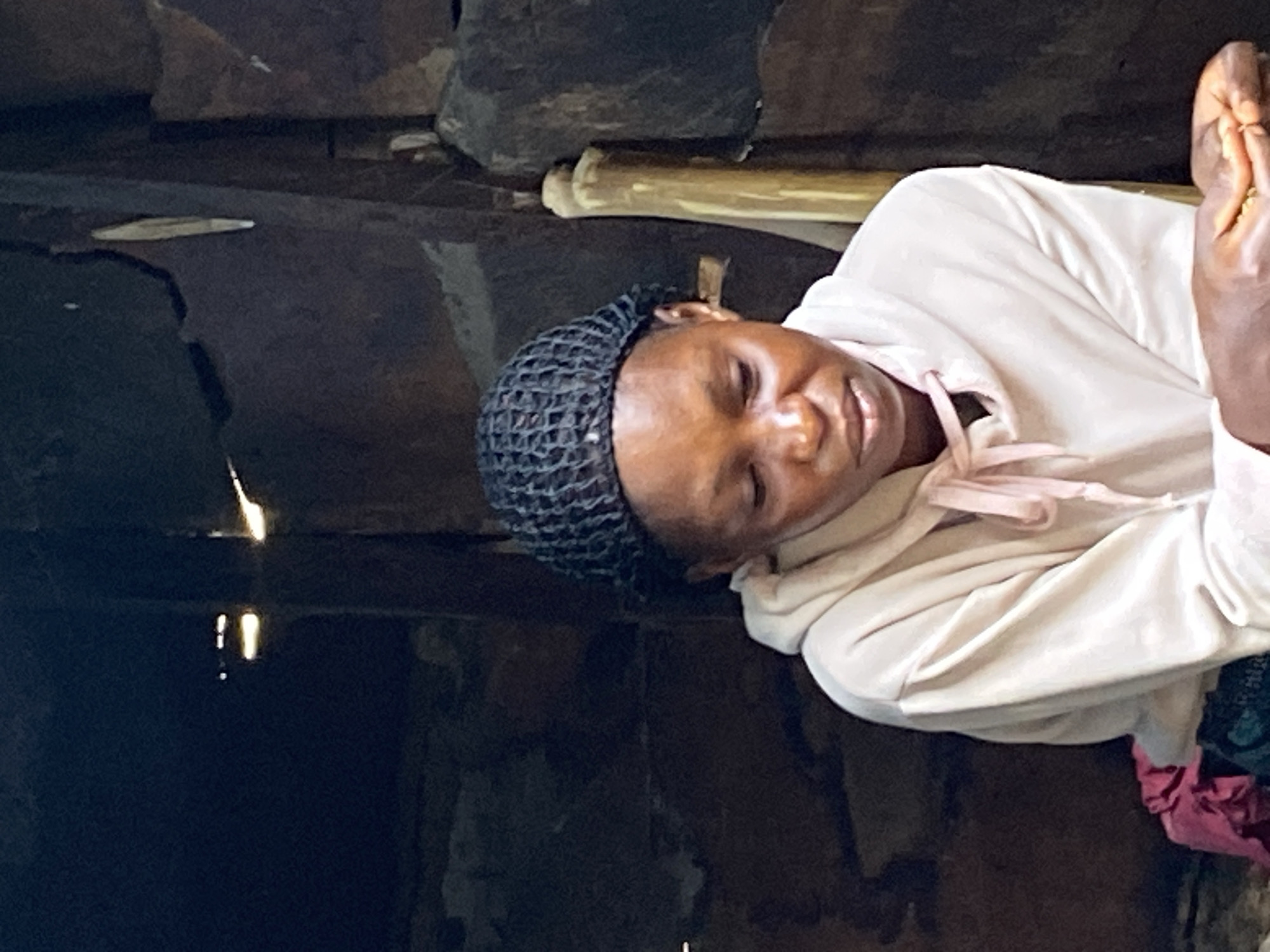 A person with dark skin and wearing a black hairnet is lying down next to a body of dark, still water. The person is wearing a light pink or white hoodie with pink drawstrings. The water reflects light, and there appears to be a bamboo-like object behind the person. The setting appears to be a shaded, enclosed area, possibly inside a structure, with dark-colored materials forming the walls or ceiling. The scene is presumed to be in Mararaba, Nigeria, based on the prompt.
