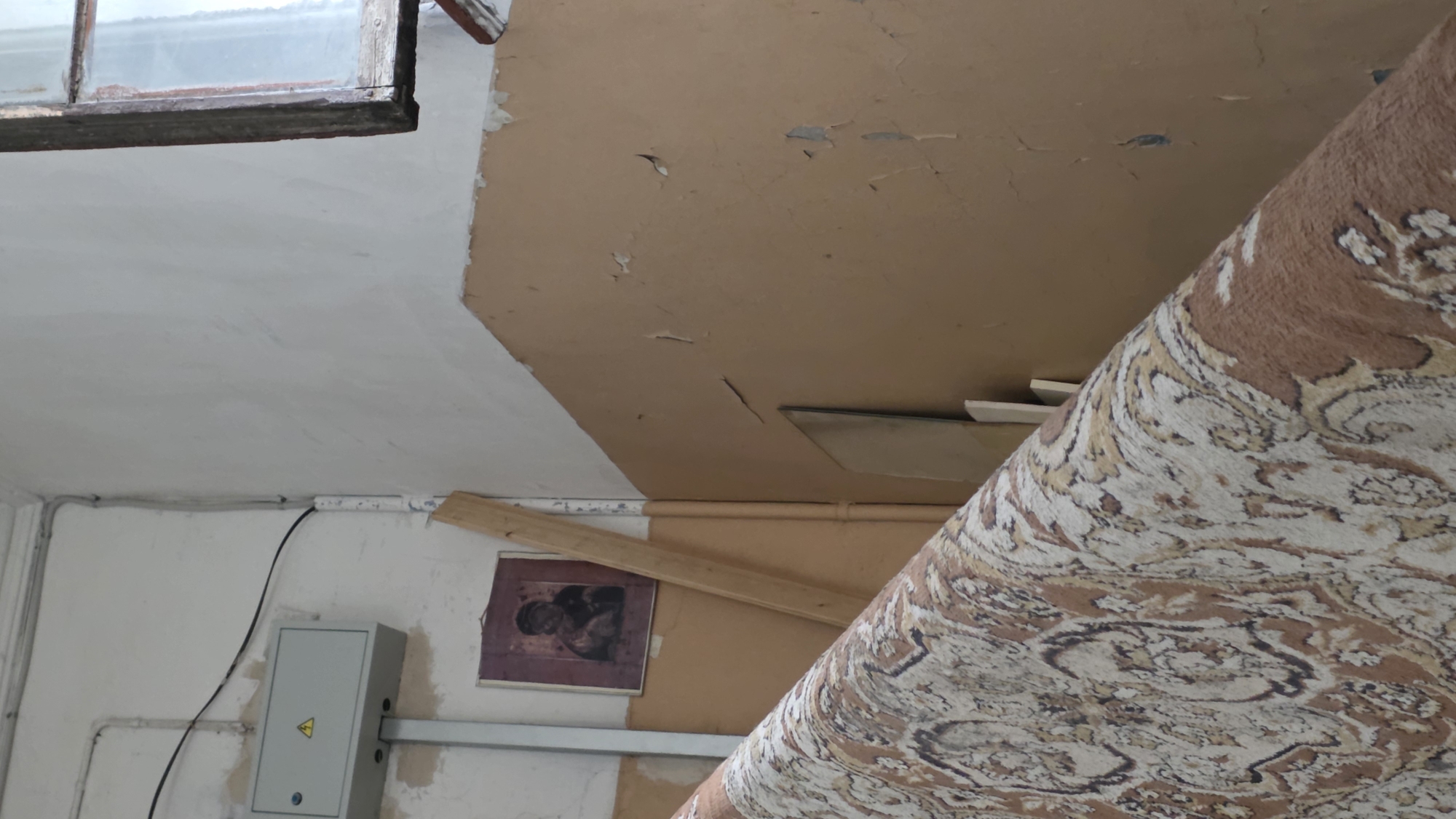 The image shows the interior of a room with damaged walls and ceilings. The upper left corner features a window. The ceiling is divided into two sections, one painted white and the other brown with peeling paint. A rolled-up carpet with a brown and white pattern takes up a large portion of the right side of the image. On the lower left, there's a grey electrical box with a yellow warning sign on a white wall. A framed picture is visible on the wall near a wooden plank. The room appears to be in disrepair. The location is Unknown, Unknown.