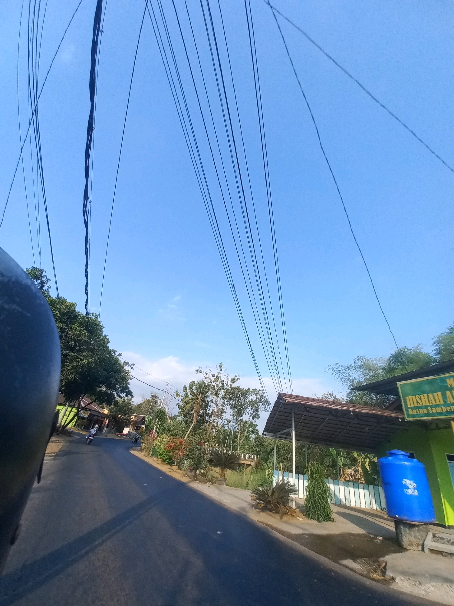 A low-angle, first-person perspective shot captures a rural Indonesian road in Kulurejo. The foreground is dominated by the dark, curved edge of a helmet, suggesting the image was taken from a motorcycle. The road itself is asphalt, appearing well-maintained, and stretches into the distance where it curves gently. Two motorcycles are visible on the road, one closer and one further away, with riders. Lush green trees line the left side of the road, with houses peeking through. On the right side, a green building with a sign that reads "HISHAR AL-Dusun Samben" is partially visible. A large, bright blue water tank stands next to the building. Overhead, a dense network of electrical wires crisscrosses the clear blue sky, creating a striking pattern. The overall scene depicts a bright, sunny day in a village setting.