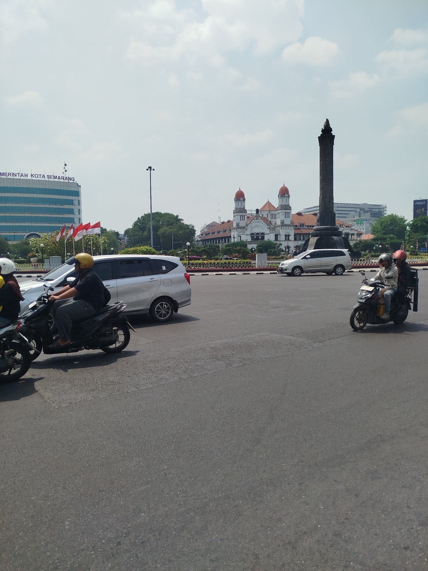 The image shows a street scene in the city of Semarang, Indonesia. In the background, the prominent Lawang Sewu building with its distinctive red-domed towers and the Sam Poo Kong temple complex are visible, alongside modern high-rise buildings and a large monument. In the foreground, several vehicles are present, including a silver minivan and multiple motorcycles. Two men on a motorcycle are in the process of turning, and another motorcycle with a rider wearing a yellow helmet is also visible. The road appears to be a paved intersection. The sky is mostly clear with scattered clouds, indicating daylight. The overall atmosphere suggests a busy urban environment.