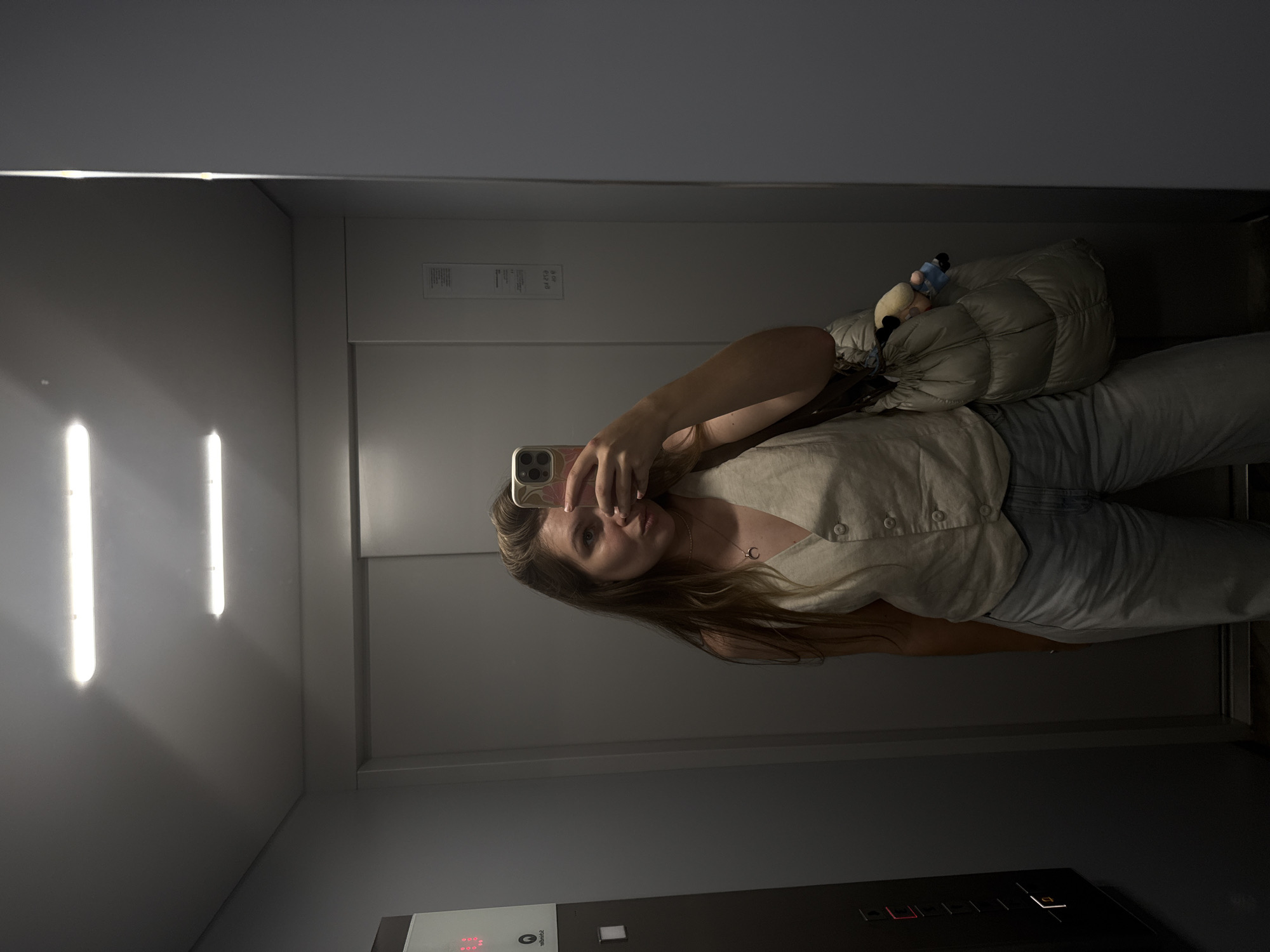 A woman is taking a selfie in what appears to be an elevator. She is holding a phone in her right hand, obscuring part of her face. She has long brown hair and is wearing a light-colored button-down shirt and light-wash jeans. A puffy bag is slung over her shoulder. The elevator has two vertical fluorescent lights on the left, with control panels below and informational panels above. The background is a plain gray. The location is within the Vozdovac Urban Municipality, Serbia.