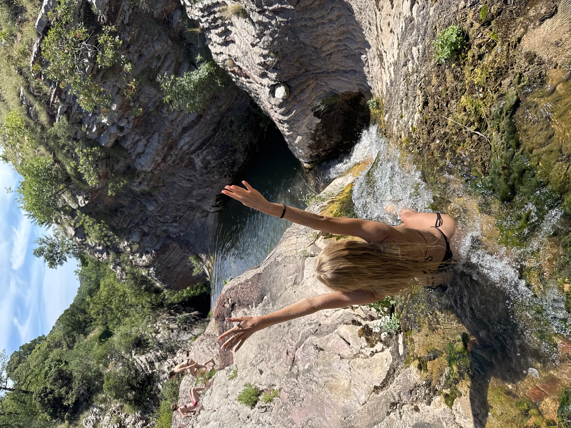 The image depicts a natural environment with rocky terrain and vegetation. In the foreground, a woman with blonde hair is sitting in shallow water at the edge of a waterfall, with her arms raised above her head. She is wearing a black bikini. In the background, two other individuals, also in swimwear, are visible on a rocky outcrop. The surroundings include lush green trees and layered rock formations. The scene suggests an outdoor recreational activity, possibly swimming or enjoying a natural waterfall. The specific location in Rafailovići, Montenegro, is not visually evident from the provided image.
