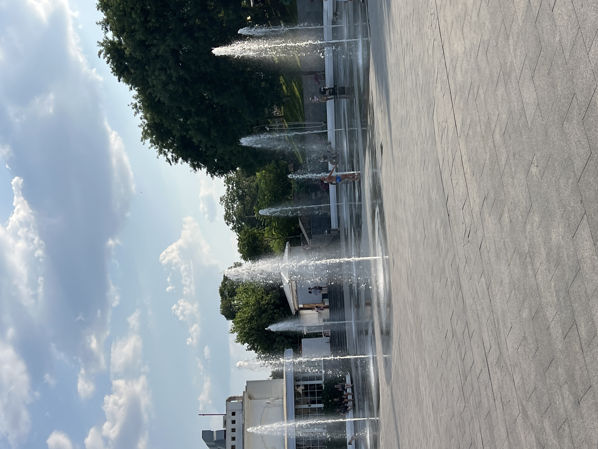 The image shows a splash pad with multiple water jets spraying upwards, creating a playful and refreshing atmosphere. Several children and adults are visible enjoying the water, with some running through the jets and others observing from the side. The setting appears to be an urban park or public square, with trees and buildings in the background. The ground is paved with grey tiles, and the water from the jets creates reflections on the wet surface. The sky is partly cloudy, suggesting a pleasant day, likely during daytime. The overall mood is joyful and summery, evoking a sense of carefree fun. There is no visible text in the image.