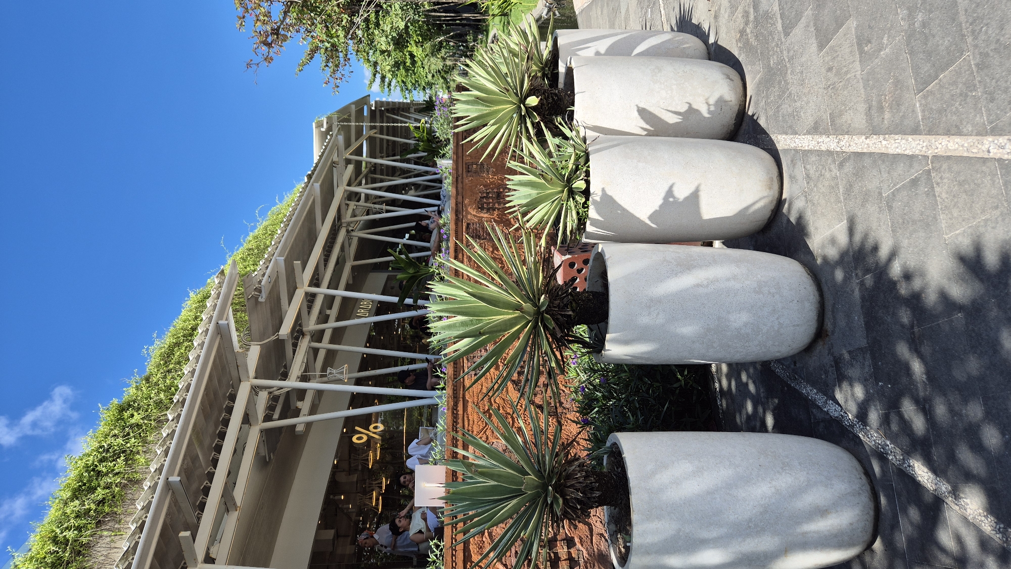 The image depicts a scene in Kuta, Indonesia. A line of four elongated, cylindrical concrete planters contains agave plants with yellow-green leaves and dark, almost black soil. The planters sit on a grey tile walkway. Behind the planters is a low, rusted-orange brick wall. Beyond that, a modern building with a white, open-frame structure is visible. Greenery covers the top edge of the building. People are inside the building, which seems to be a cafe or restaurant, with signs and logos visible on the interior. The sky is clear blue. Shadows from nearby trees fall across the walkway.