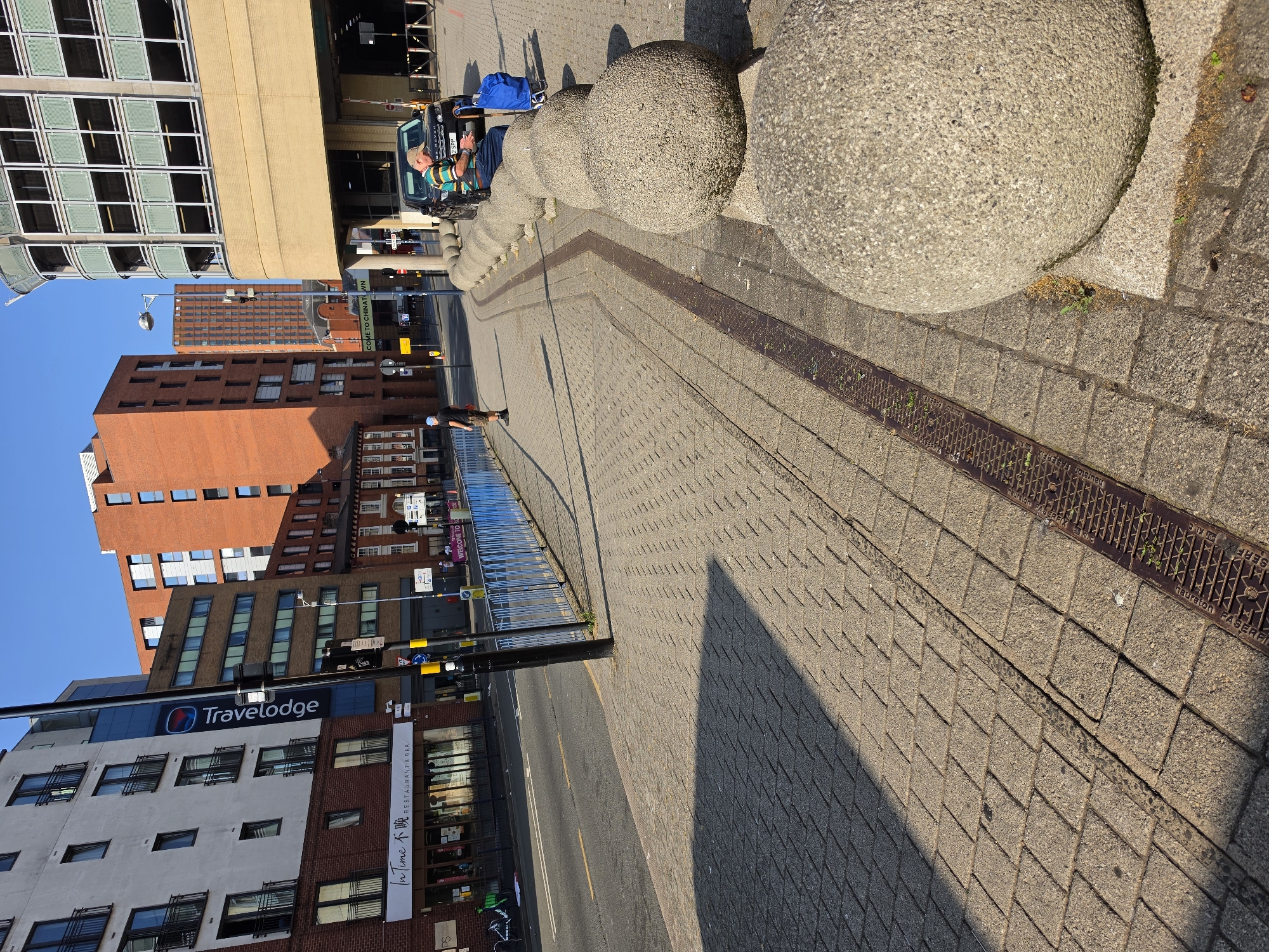 The image depicts a street scene in Birmingham, United Kingdom, likely near the Chinatown area, given the "Welcome to Chinatown" sign visible in the background. The perspective is from a sidewalk paved with textured concrete tiles, casting a long shadow from an unseen structure to the left. A metal drain grate runs parallel to the sidewalk's edge, with small weeds growing through it.
Along the right side of the sidewalk are large, spherical concrete bollards, placed in a line. One of these bollards is in the foreground, partially obscuring the view of the sidewalk beyond. Behind the bollards, a dark-colored SUV is parked. A man wearing a striped polo shirt, dark pants, and a baseball cap is seated on the bollards next to the SUV, looking at his phone. A blue shopping bag with wheels is beside him.
Further down the sidewalk, a person in a dark blue t-shirt and dark shorts is walking away from the camera. A bright blue metal fence runs along the street separating the sidewalk from a road. On the left side of the road, a multi-story building with white and red brick facades is visible, featuring numerous windows and balconies. A sign for "Travelodge" is prominently displayed on this building. Other buildings of varying architectural styles and heights are present in the background, including a tall, orange-brick building. The sky is clear and blue.