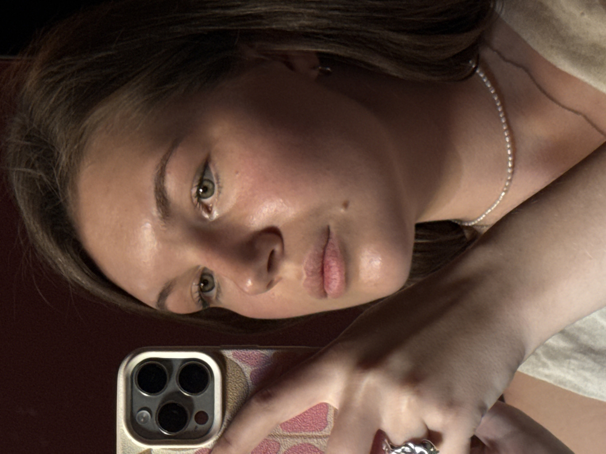 A close-up selfie of a Caucasian young woman is captured with an iPhone. The woman's face is angled towards the camera, displaying light brown hair, fair skin, and light-colored eyes. She has a mole on her right cheek. She is wearing a delicate silver necklace. Her right hand is holding the phone, with a ring visible on her finger. The phone case features a beige and pink pattern. The background is out of focus, appearing as a dark, indistinct red. The image does not provide information to identify the specific location in Belgrade, Serbia. There is no information on any interactions.