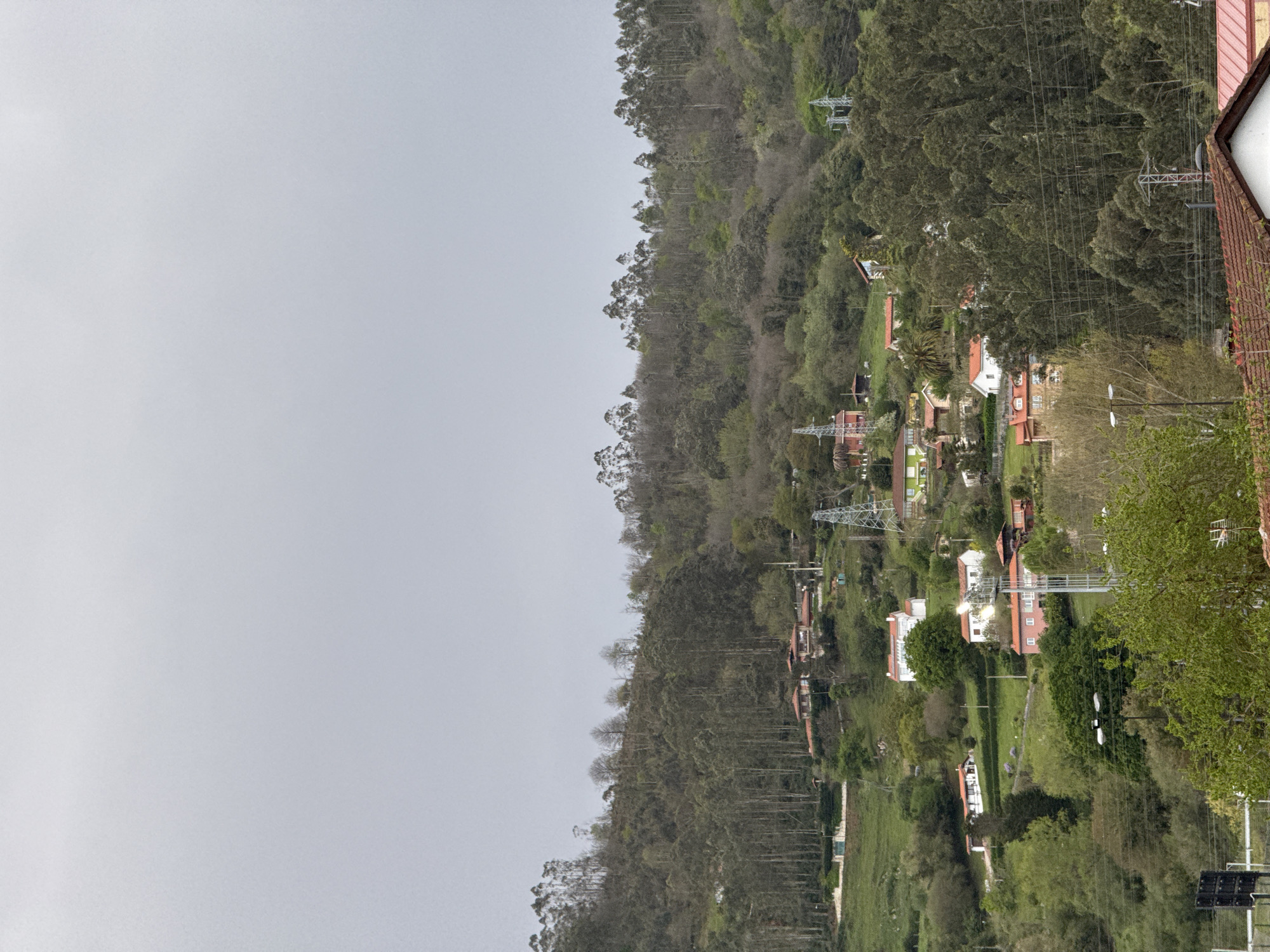 The image depicts a hillside residential area in El Pontón De Vaqueros, Spain. The hillside is covered with trees of varying heights. Homes are scattered throughout, with a mix of red-tiled roofs and white walls. Power lines and electrical towers are visible amidst the buildings and trees. The left side of the image shows a cloudy sky. The hillside is heavily forested.
