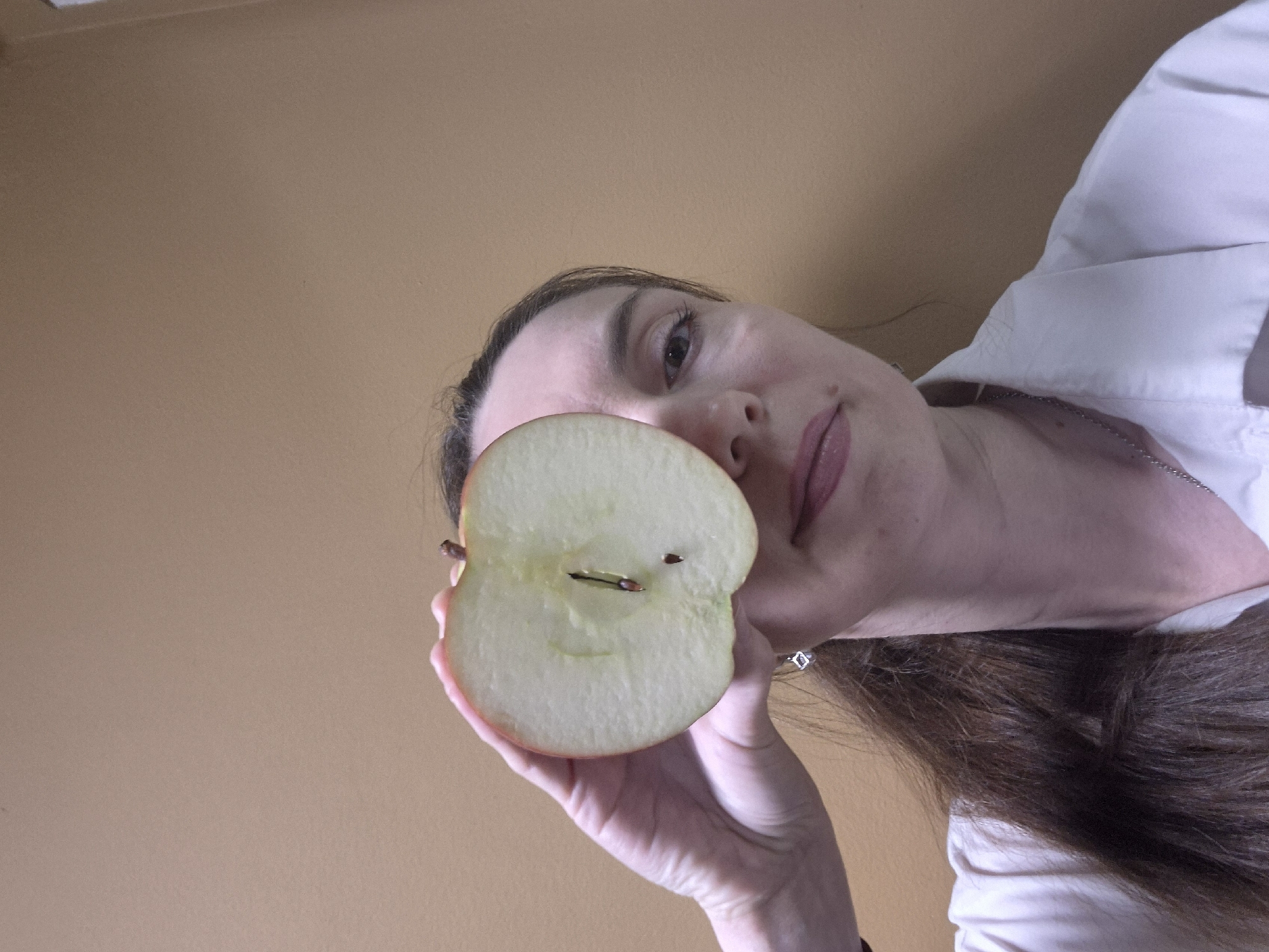 The image captures an adult female from a low-angle perspective, looking upwards. Her face is partially visible, with her right eye looking directly at the viewer, and her lips forming a subtle smile or smirk. She has long, dark brown hair falling across her shoulder and chest. She is wearing a light-colored, possibly white, collared shirt, with a thin silver chain visible around her neck.
In her right hand, held prominently towards the camera, is a red apple cut vertically in half. The apple's white flesh is clearly visible, along with multiple dark seeds; one is centered within the core, and another smaller seed is closer to the edge. A small piece of the apple stem protrudes from the top part of the cut surface. On her right ring finger, she wears a silver ring featuring a clear, rectangular or square stone.
The background consists of a plain, light brown or beige wall, suggesting an indoor setting. The lighting is diffused and even, with no strong shadows to indicate a specific time of day. No text is visible in the image. The overall impression is a direct, somewhat enigmatic portrait with the cut apple as a focal point.