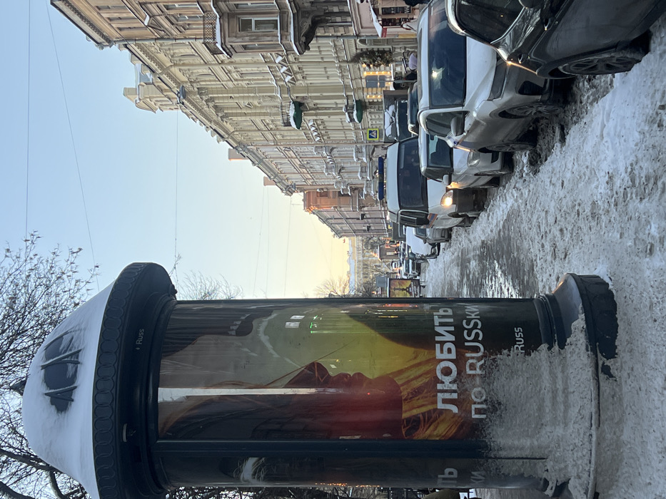The image depicts a winter street scene in Saint Petersburg, Russia, specifically Nevsky Prospect, looking towards the Kazan Cathedral in the distance. The environment is characterized by clear skies and snow-covered ground.
In the foreground on the left, a dark grey cylindrical advertising column stands partially covered at its base and top with slushy snow. The column displays an advertisement featuring two silhouetted profiles, possibly of a man and a woman, rendered in warm red and yellow tones. Below the profiles, Cyrillic text is visible, reading "ЛЮБИТЬ по-RUSSKI" ("To love in Russian"). The word "Russ" is also present as a logo.
The sidewalk adjacent to the column and extending into the midground is unevenly covered with compacted snow and slush, showing signs of foot and tire traffic.
On the right side of the street, a line of vehicles is either parked or slowly moving. Closest to the camera is the rear of a dark-colored SUV, followed by a lighter-colored SUV. Further down the street, a white cargo van with blue lights on its roof, potentially a police or utility vehicle, has its headlights illuminated. Other sedans are visible in the line of traffic. The street itself is covered in snow and slush, particularly where vehicles are parked.
Lining the right side of the street is a row of multi-story classical buildings, characterized by light-colored facades (beige, cream), ornate architectural details, and snow accumulation on cornices and ledges.
In the far background, a large, grand classical building with columns and a prominent central structure, identified as the Kazan Cathedral, is visible, illuminated by the low-angle sunlight which casts a warm, yellowish hue on the horizon. Bare tree branches are discernible against the clear blue sky on the upper left. No discernible individuals are performing actions or interactions within the frame.