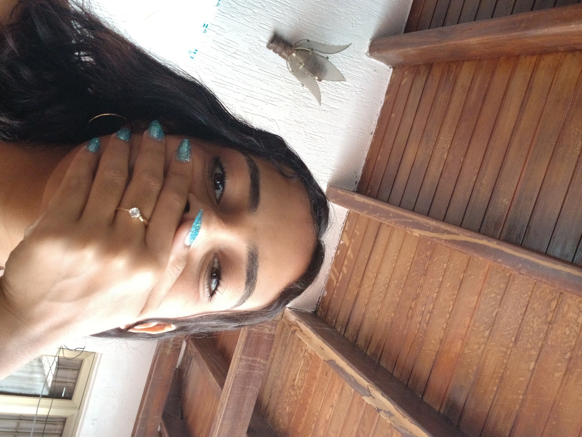 The image is a selfie, seemingly taken from a low angle, featuring a woman indoors. Her face and hand are prominently displayed. She has long, dark hair, and is wearing a ring on her ring finger. Her fingernails are painted with glittery blue polish. Her hand is partially covering her mouth.
The background includes a white ceiling with a decorative light fixture, and wooden planks resembling a ceiling or roof structure. Part of a window with closed blinds can be seen in the corner.
The location, Juan Griego, Venezuela, is not visually verifiable in this image. Without further context, it is impossible to ascertain the location based on the visual information present.