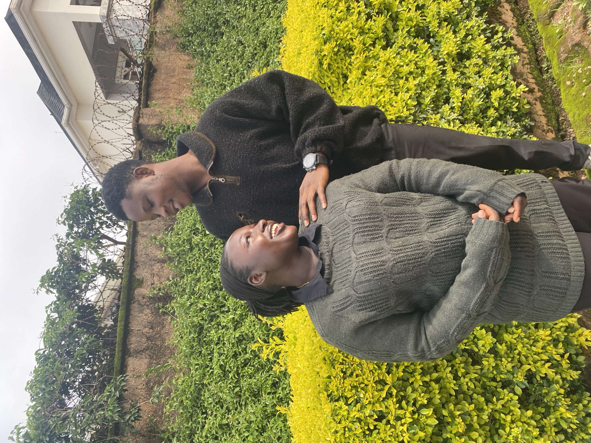 This media file shows a close-up, vertical shot of a young Black couple outdoors, with a building and lush green foliage in the background. The man, on the left, is wearing a black fleece-like sweater and looking down at the woman with a gentle smile. He has short black hair and a watch on his left wrist. The woman, on the right, is wearing a dark grey cable-knit sweater and looking up at him, laughing heartily with her mouth open. She has dark hair pulled back and appears to be wearing a dark head covering.
The couple is positioned in front of a dense, vibrant green hedge that transitions into a brighter yellow-green shrubbery. Behind them, to the left, a light-colored building with multiple levels and windows is visible, topped with a dark roof. Barbed wire runs along a textured wall in front of the building, suggesting a sense of security or enclosure. The overall impression is of a warm, affectionate moment shared between the couple in a semi-urban or suburban setting. The lighting suggests it's daytime, likely overcast given the diffused light. The setting, combined with the mention of Jos, Nigeria, places the scene in this specific geographical context. The emotions conveyed are joy and intimacy. There is no visible text in the image.