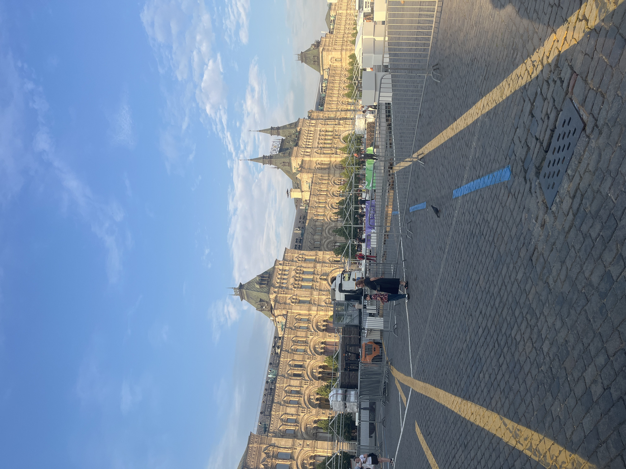 The image depicts a wide cobblestone plaza in front of a large, ornate building with a distinctive architectural style, characteristic of Moscow, Russia. The building, which appears to be the State Department Store (GUM) on Red Square, features a long facade with numerous arched windows and multiple towers topped with green spires. The sky is a clear blue with scattered white clouds.
In the foreground, the cobblestone pavement is marked with yellow lines, suggesting a designated area or pathway. Several metal barriers are erected, creating a temporary enclosure or perimeter. Within this enclosure, there are various objects that indicate preparation for an event or setup. These include white tents or canopies, stacked materials that appear to be pallets of goods, a forklift, and a white truck.
Several individuals are present in the plaza. Two women and a child are standing near the barriers. One woman is wearing a dark dress and the other a white t-shirt and shorts. A child in a patterned shirt stands next to one of the women. Further back, more people are visible, some working or observing the setup. A lone pigeon is seen walking on the cobblestones. The lighting suggests it is either early morning or late afternoon, with a warm glow illuminating the scene.