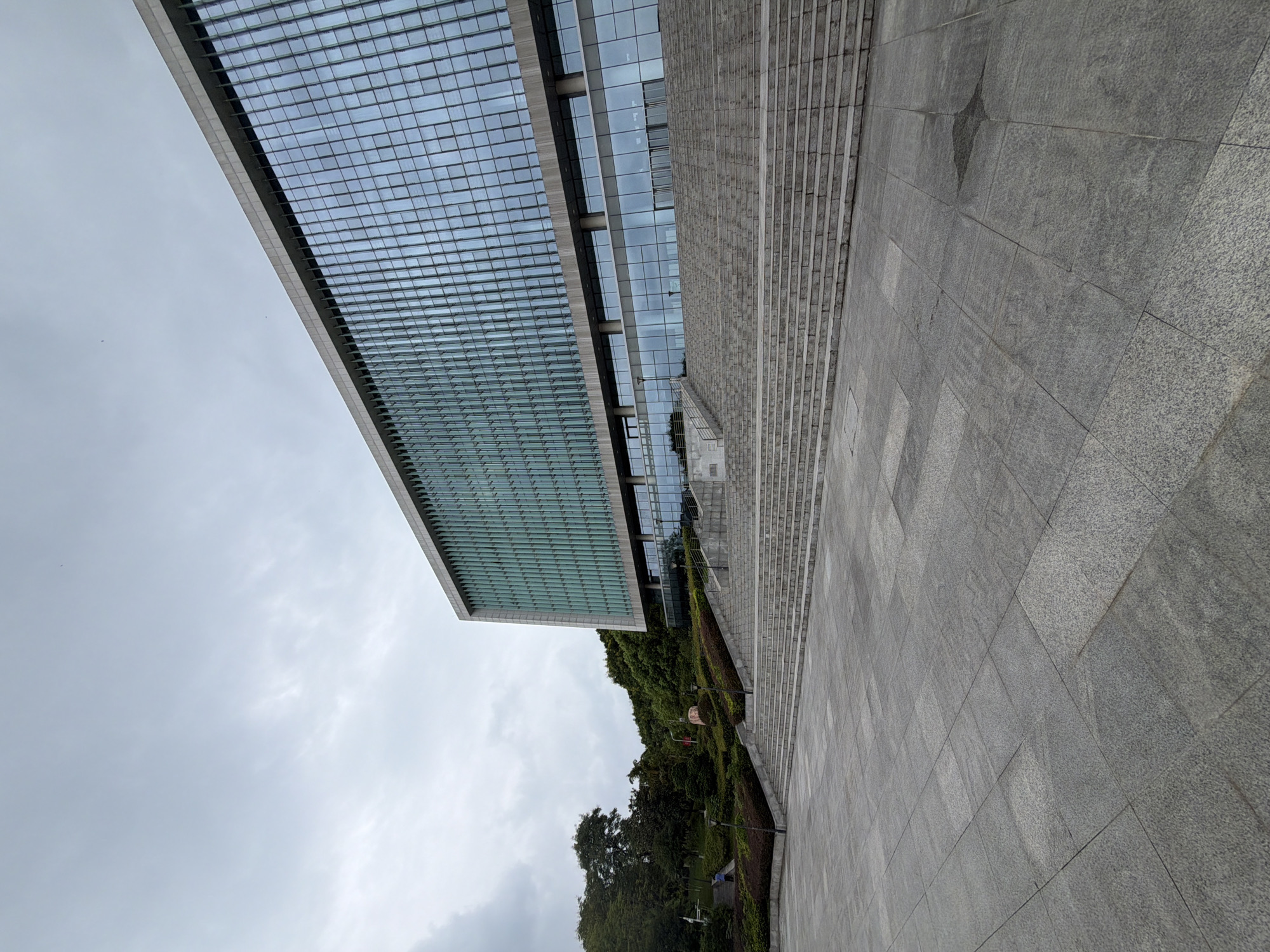 The image shows a wide, paved plaza in front of a large, modern building. The building has a facade of blue-tinted glass windows and is set against a backdrop of green hills and trees. A long, wide staircase leads up from the plaza to the entrance of the building. The sky is overcast and gray. A single person in a blue shirt is walking on a path to the left of the stairs, and another person is visible in the distance on the same path. The plaza is paved with large, gray granite tiles. This scene is located in the city of Jiangjin, China.
