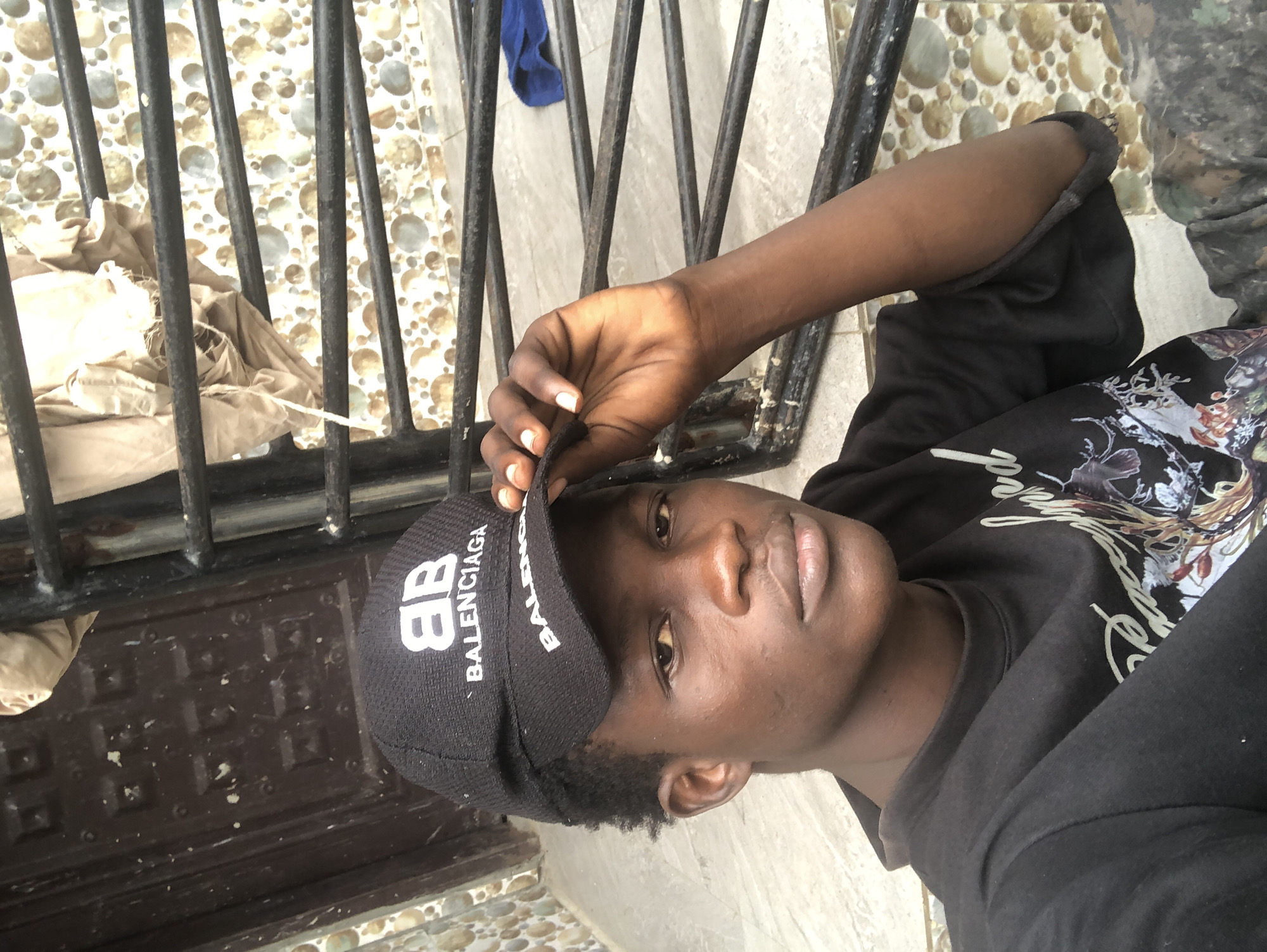 A young Black male wearing a black Balenciaga baseball cap and a black sweatshirt is posing for a selfie outdoors. He is wearing camouflage pants. The background features a brown wooden door, a tiled wall with a circular pattern, and a black metal grate or railing. A tan cloth is draped over the railing. The setting appears to be in Ado, Nigeria.