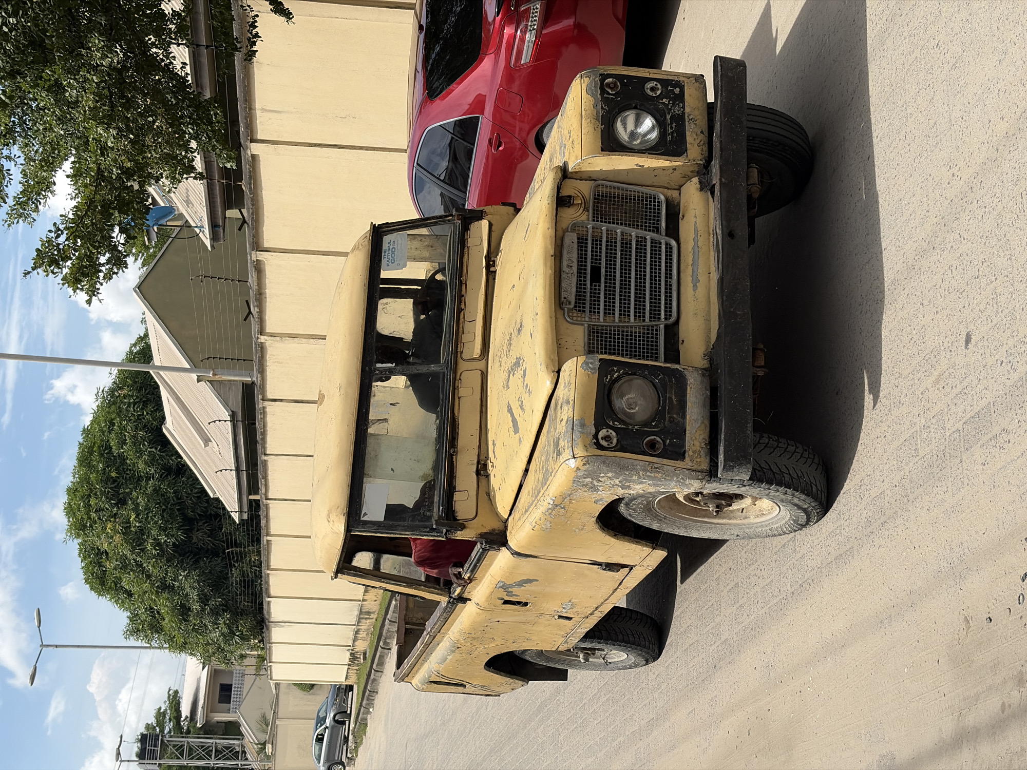 A medium shot captures a yellow, older model Land Rover parked on a paved street in Igboefon, Nigeria. The car is facing forward, with a red SUV parked closely behind it. The Land Rover exhibits signs of age and wear. A person is partially visible inside the Land Rover, wearing a red shirt. The background includes residential buildings with beige walls, green trees, and a blue sky. Streetlights and other parked cars are visible further down the street.