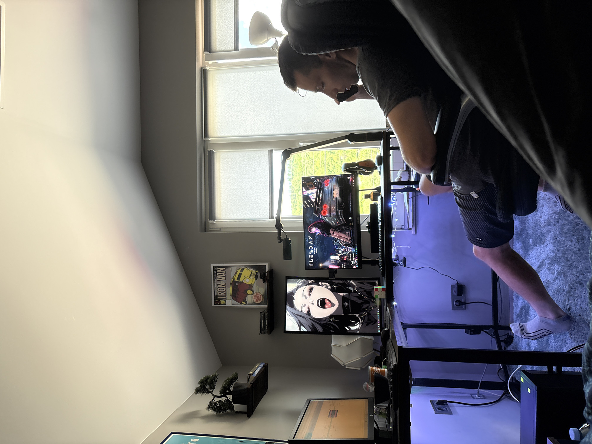 A man sits in a gaming chair in a room, speaking on a black mobile phone. He is wearing a dark grey t-shirt and dark shorts. The room is dimly lit, with blue LED strip lights illuminating the underside of a desk. The desk has three computer monitors, keyboards, and various other items. One monitor displays an anime image of a woman with her mouth open. Another monitor displays a car. The third monitor displays what appears to be a spreadsheet or code. On the wall above the desk, there are two framed posters and a small artificial bonsai tree on a shelf. Large windows with blinds cover one wall of the room, offering a view of greenery outside. The floor is covered in a thick, light-colored rug. The image appears to be taken indoors in a residential setting, possibly a bedroom or office, in Gabrovo, Bulgaria.