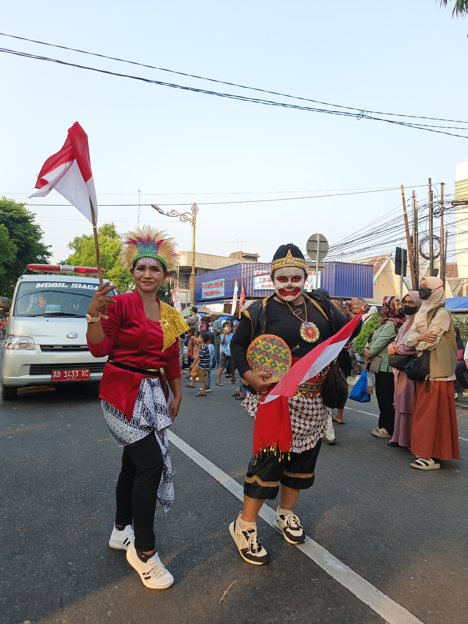 In the city of Klaten, Indonesia, two individuals in traditional attire are participating in an outdoor event, possibly a parade. The person on the left is a woman with her face painted and adorned with a colorful feathered headdress. She wears a red top and a white patterned sarong, holding an Indonesian flag and making a peace sign. To her right, a person in full traditional makeup and costume, including a crown, holds a fan and an Indonesian flag. They wear a black top with ornate trim and a patterned sarong. Behind them, a silver van with a license plate reading "AD 1433 XC" is visible. The background shows a street lined with buildings, trees, and utility poles, with other people observing the event. The ground is asphalt, marked with a white line.