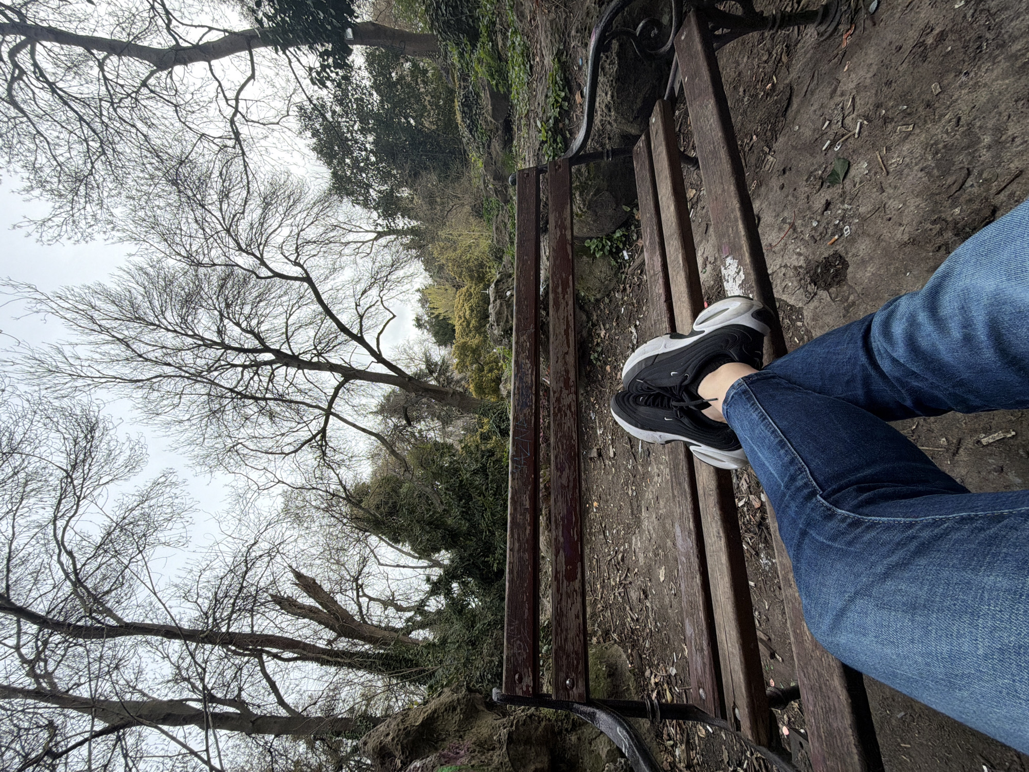 A person wearing blue jeans and black/white sneakers is sitting on a wooden park bench. The bench is located in a wooded area with bare trees and some green foliage visible in the background. The ground around the bench is covered in dirt, leaves, and litter. The scene is possibly located in Varna, Bulgaria, in one of the parks or green spaces within the city.