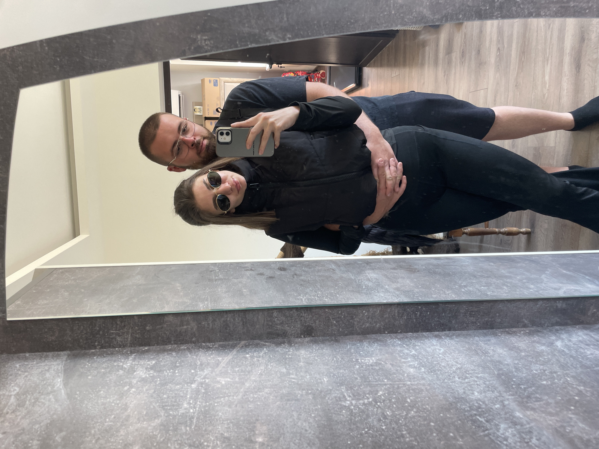 A couple poses for a selfie in a mirror. The man has short hair, a beard, and glasses, wearing a black t-shirt and shorts. The woman wears sunglasses and a black vest over a black top with black pants. The man's arm is around the woman, and she holds the phone, taking the picture. The background features a wooden floor, cream-colored walls, and glimpses of furniture and objects, including a cardboard box. The location cannot be determined from this image alone.