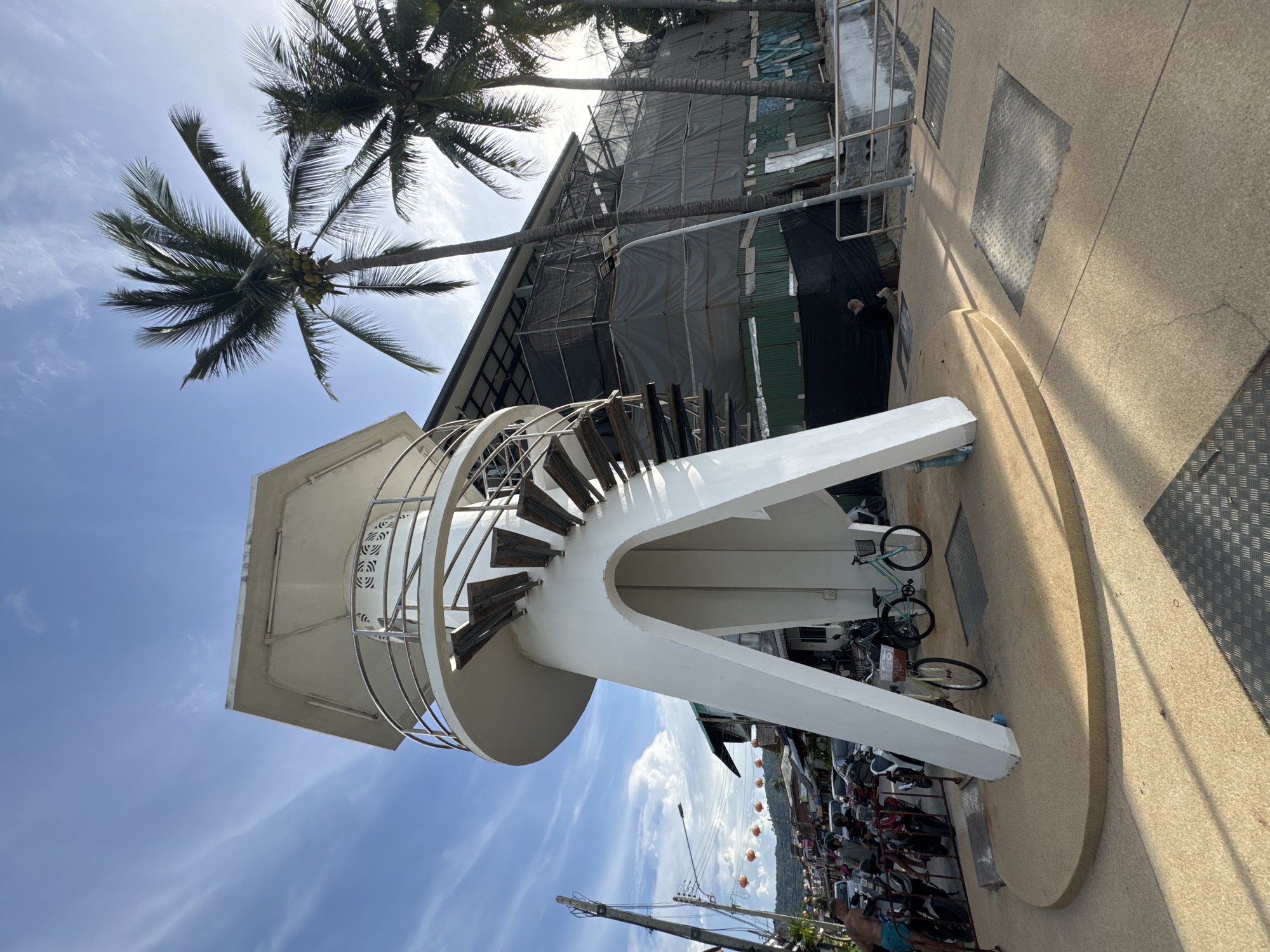 A bright, sunny day in Ko Samui, Thailand, is captured in this image, with the sky a vivid blue dotted with wispy clouds. The scene appears to be a coastal area, likely a tourist spot given the presence of parked motorcycles and bicycles.
The focal point is a modern, white spiral staircase that ascends around a cylindrical structure. The staircase features wooden steps and a sleek metal railing. The base of the structure is made of rough, sandy-colored concrete, with what looks like metal grates embedded in it. Behind the staircase, a building is under construction, covered in grey and green tarps, with scaffolding visible. Palm trees with lush green fronds are scattered throughout the frame, adding to the tropical ambiance.
In the foreground, several bicycles are parked near the base of the staircase. To the right, a line of motorcycles is visible, suggesting a busy area. A person, partially visible in dark clothing, stands near the construction site. Further in the background, a street scene unfolds with more motorcycles and what appear to be red lanterns strung up, indicating a festive or commercial area.
The lighting suggests it is daytime, possibly late morning or early afternoon, given the strong shadows cast by the staircase and the bright sunlight. The overall mood is vibrant and active, indicative of a lively tropical location. No visible text is present in the image.