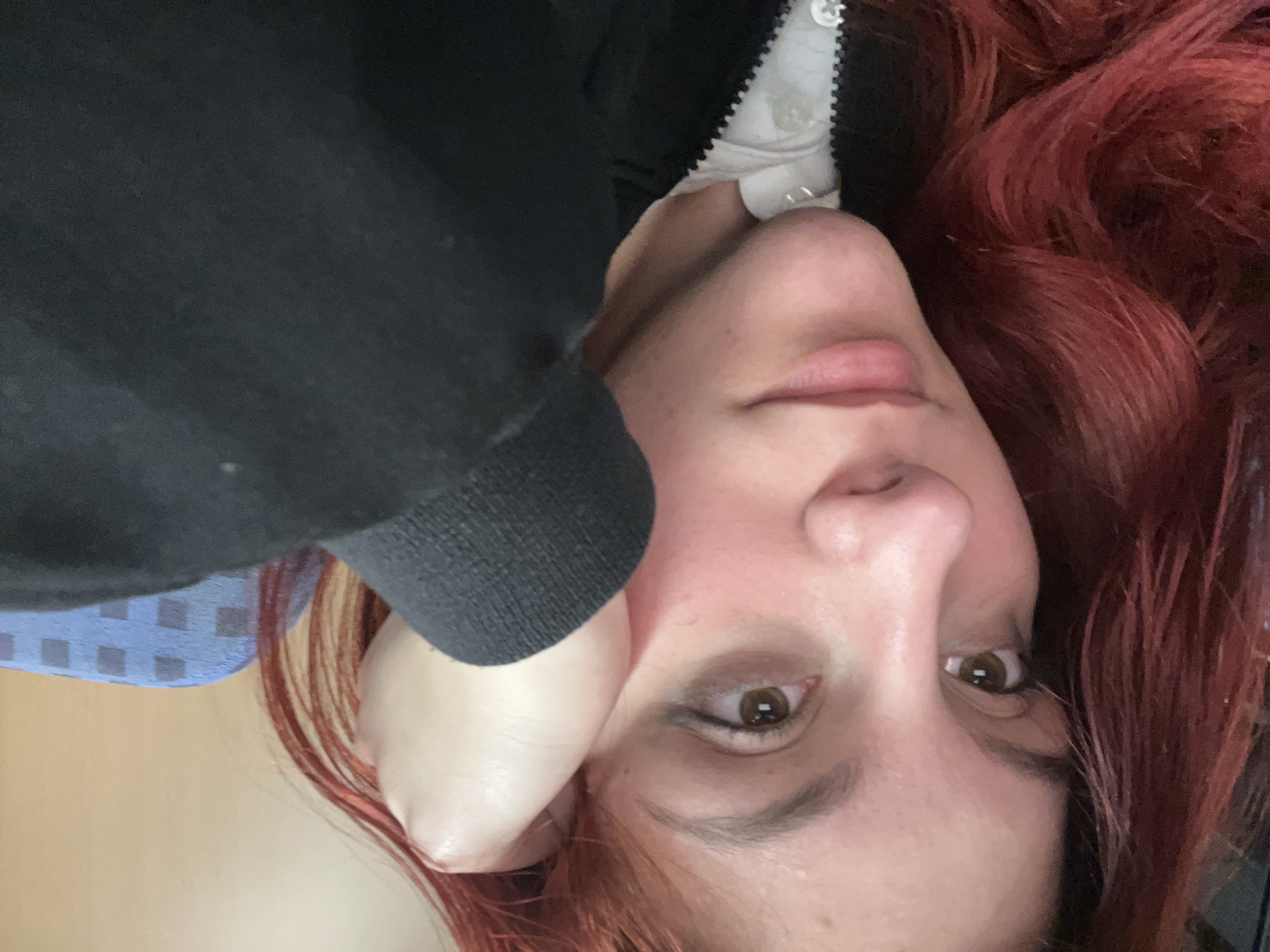 A close-up selfie of a woman with red hair. She is wearing a black jacket with a white shirt underneath. Her left hand is resting against her cheek. The background appears to be a blue and white patterned fabric, and a light wood surface. Location: unable to determine based on the provided image. The location could not be confirmed as Krumovo, Bulgaria.