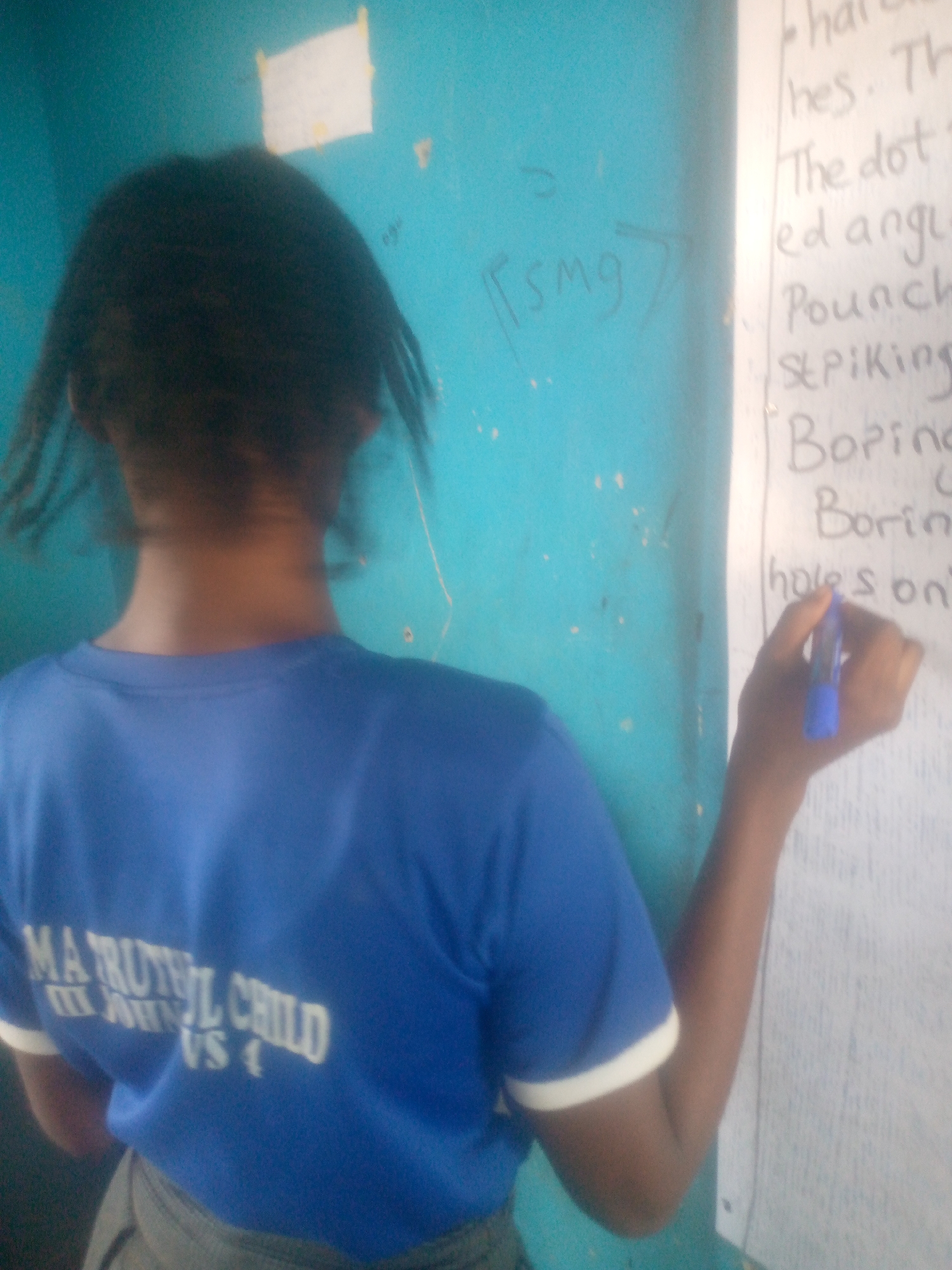 A person stands facing a whiteboard, writing with a blue marker, possibly in a classroom setting. The person is wearing a blue t-shirt with white lettering "MA TRUTIFUL CHILD" and white trim on the sleeves. The environment appears to be in Ikorodu, Nigeria, based on the lettering on the t-shirt. The whiteboard contains lists of words and sentences. The wall behind the person is painted blue.