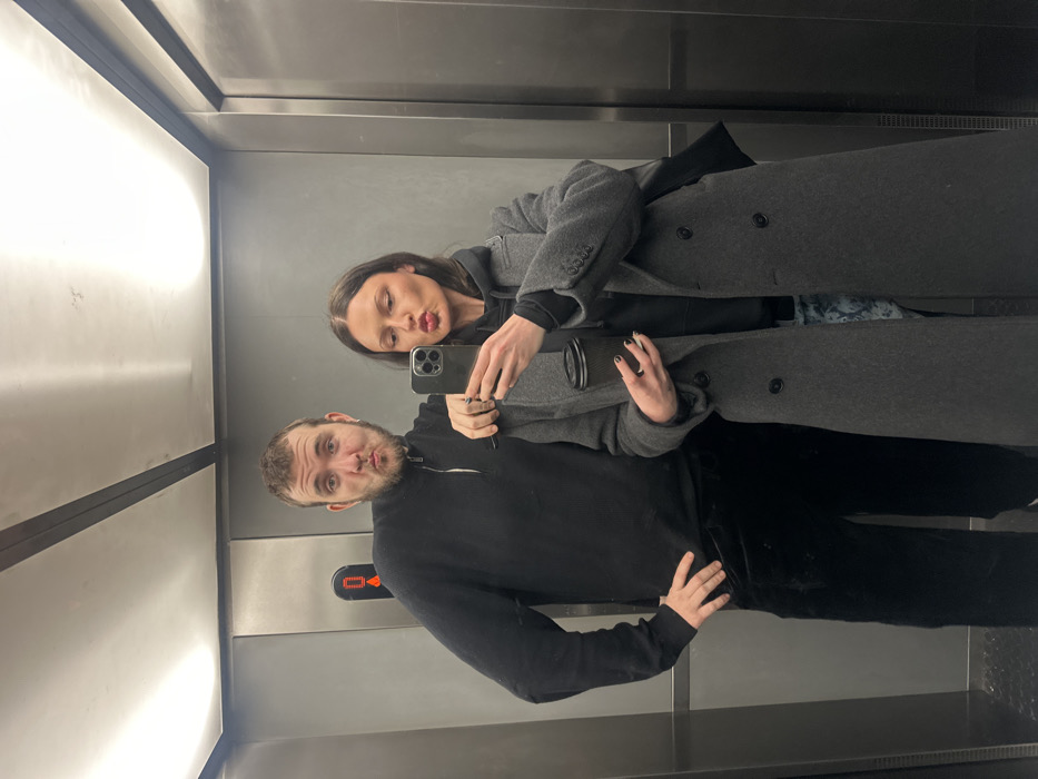 An elevator mirror selfie depicts two individuals, one male and one female. The male, positioned on the left, has short brown hair and a beard, wearing a black ribbed quarter-zip sweater and dark velvet trousers. His left hand is on his hip, and his right arm rests on the female's left shoulder. He has a small earring in his left ear. The female, on the right, has long dark brown hair parted in the middle, wearing a dark grey long overcoat over a black hooded sweatshirt and patterned trousers. She has dark nail polish on both hands. She holds a dark smartphone in her right hand, angled to capture the reflection, and a black disposable coffee cup in her left hand. Both individuals have pursed lips, forming a "duck face" expression. The elevator interior features metallic grey walls, linear ceiling lighting, and a control panel with a red digital display on the left wall. No specific identifying markers are present within the image to pinpoint a precise location in London, United Kingdom.