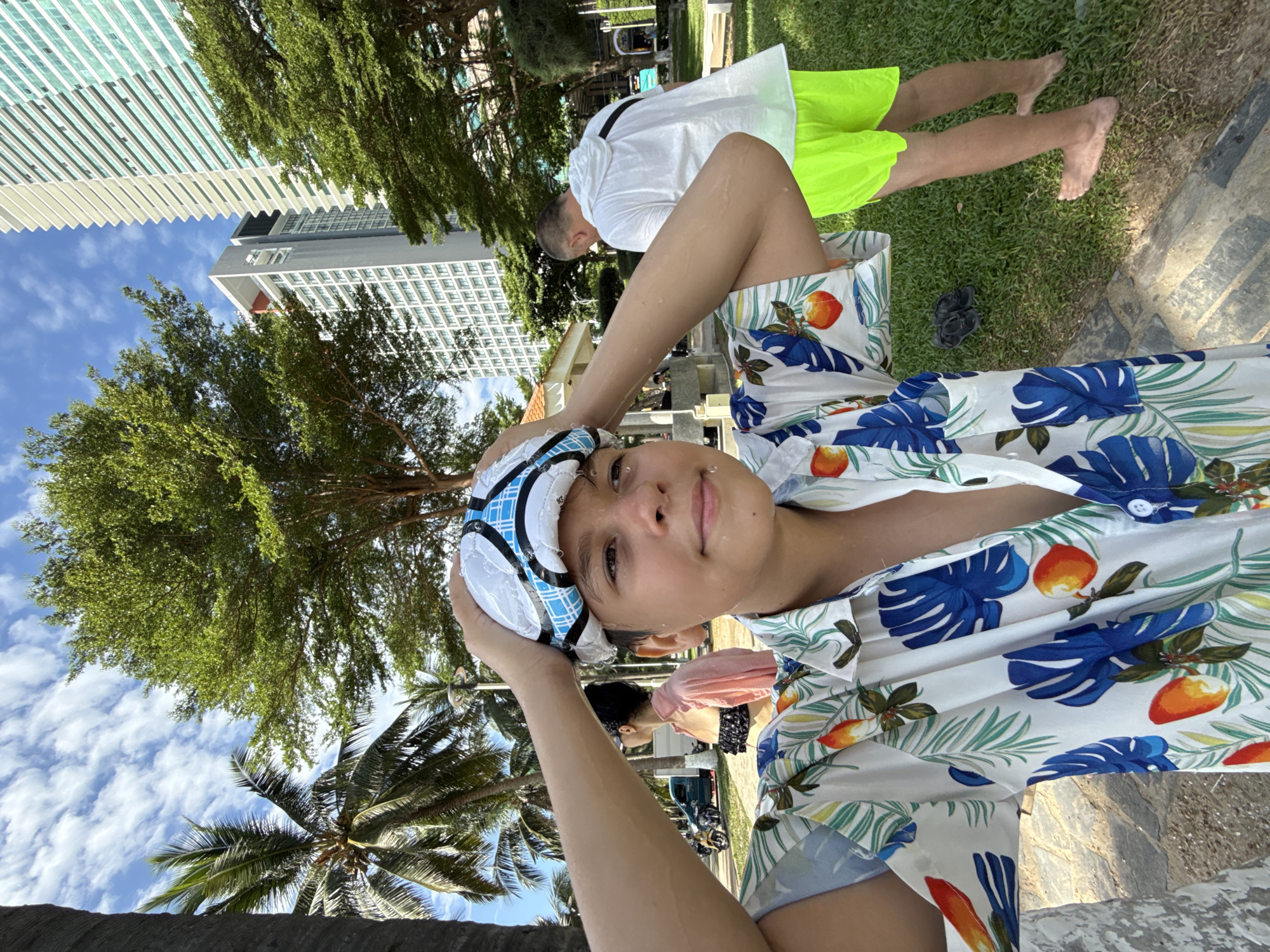 A young person is holding a soccer ball on their head, with their hands resting on it. They are wearing a short-sleeved button-up shirt with a tropical print of blue monstera leaves and orange fruit. The person's skin appears wet, suggesting they may have been swimming or are in a humid environment. Their expression is neutral.
In the background, another person is visible from the waist down, wearing bright neon green shorts and a white shirt. They are sitting on a grassy area. The scene appears to be outdoors, with palm trees and other foliage present, hinting at a tropical location. Tall modern buildings with many windows are visible in the upper left portion of the image, suggesting an urban or resort setting. The sky is blue with scattered white clouds. A patch of stone or concrete walkway is visible in the lower right.
The overall impression is a casual, warm-weather scene, possibly a vacation or leisure activity in Nha Trang, Vietnam, given the context. The time of day appears to be daytime, with clear skies and sunlight casting shadows. The presence of a soccer ball and the wet appearance of the main subject suggest a playful or athletic activity.