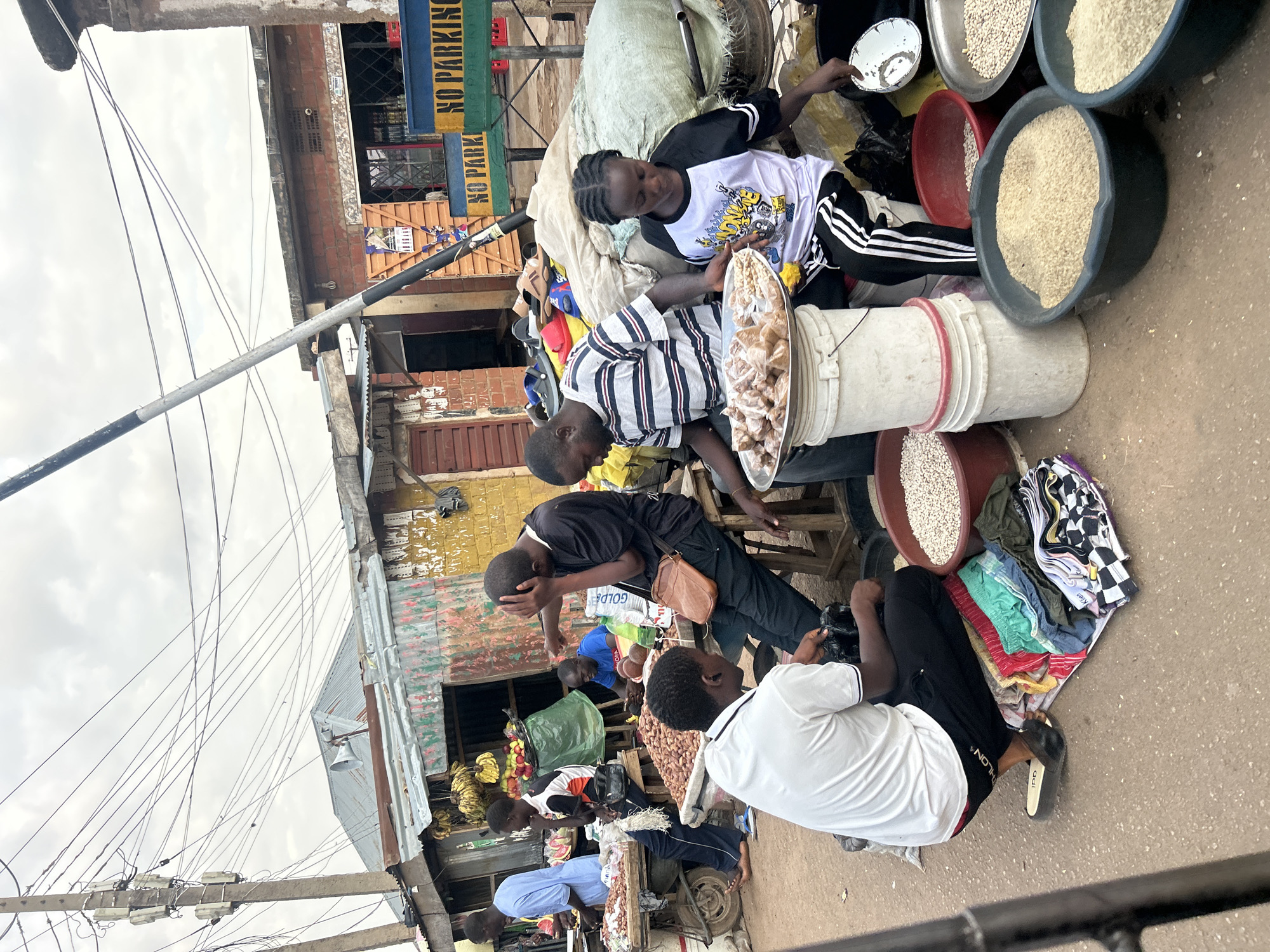 The image depicts a market scene in the city of Jos, Nigeria. Multiple people are visible. Two young vendors are seated, selling peanuts from a large container. A third vendor sits nearby, with buckets of grains around him. Other vendors are present, selling fruit and other goods. Overhead, there is a cluttered arrangement of utility poles and wires. Buildings flank the sides of the road. One building has a sign that reads "NO PARKING".