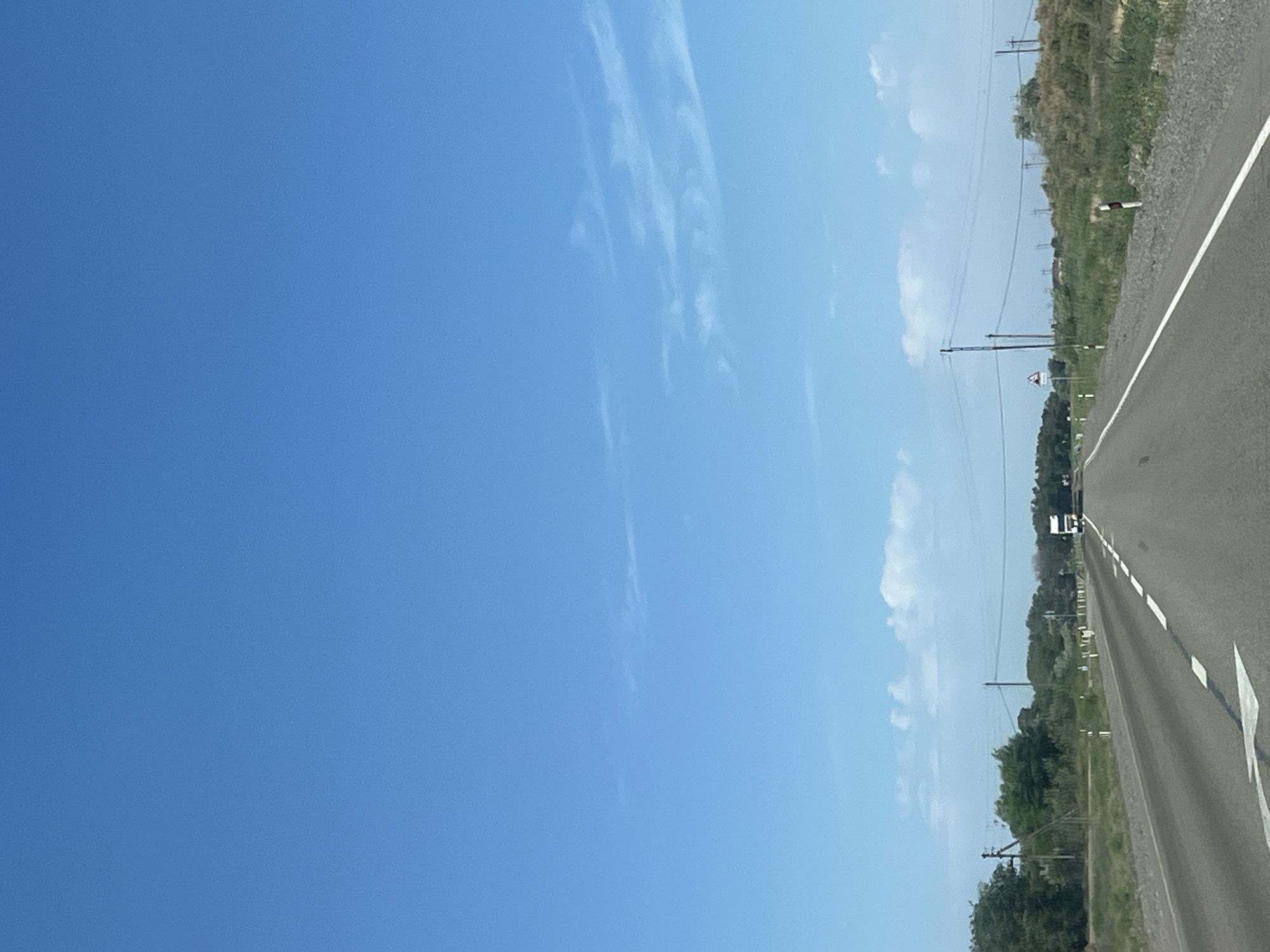 A wide-angle, forward-facing shot shows a paved road stretching into the distance under a clear blue sky with sparse white clouds. The road is marked with white dashed lines and a solid white line along the right edge. To the left of the road, there is a grassy verge with scattered trees and bushes, as well as a white arrow painted on the road surface. Further on the left, a power line pole is visible. The right side of the road features a similar grassy verge with more dense vegetation and several white and red striped road markers. In the distance, a white truck is visible on the road. There are more power line poles and wires stretching across the scene. The overall impression is a rural roadside scene in Russia, potentially near Bakhtemir, under fair weather conditions.