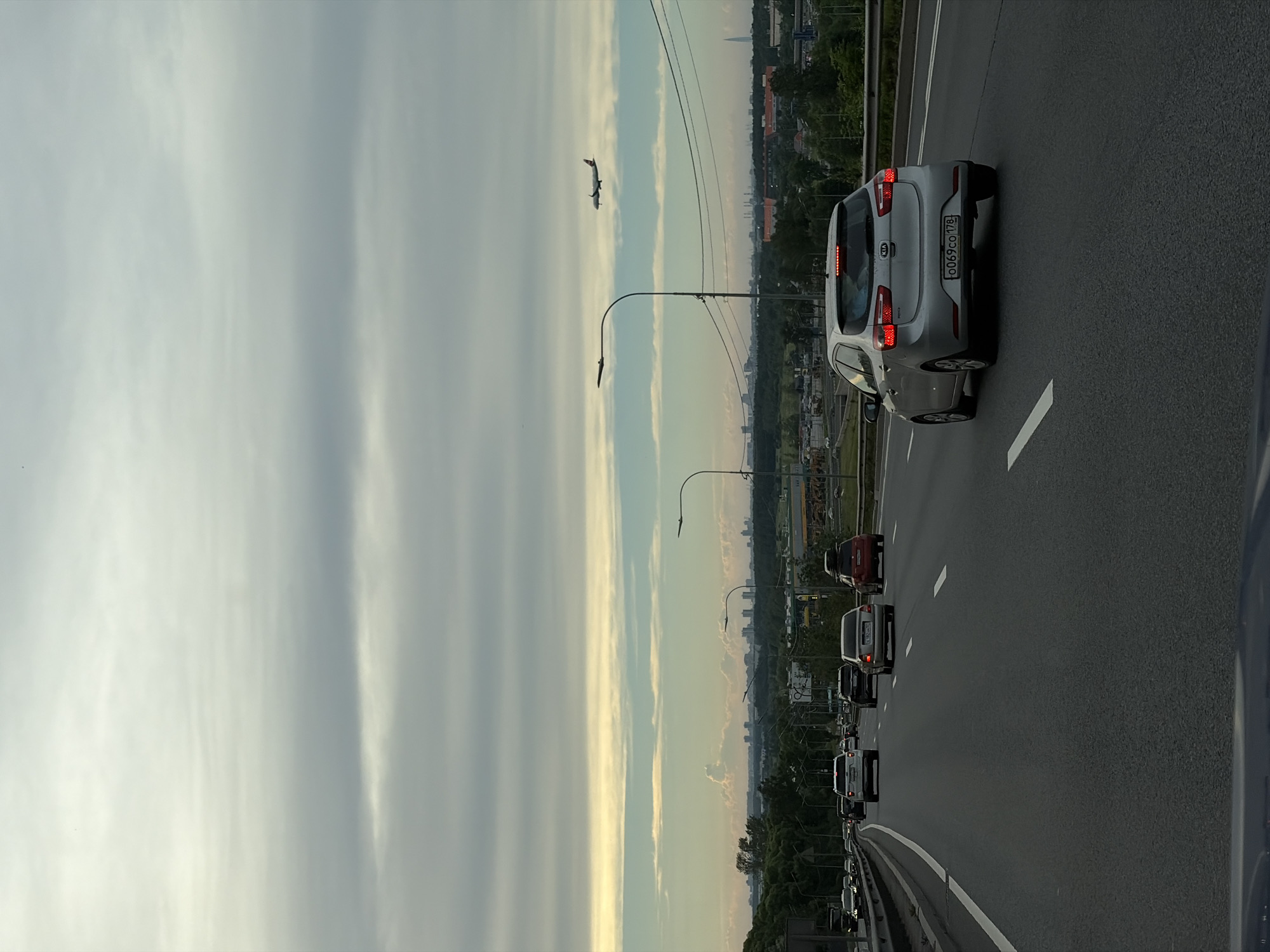 The image depicts a view from a vehicle on a multi-lane highway, likely in or near Saint Petersburg, Russia. The sky is overcast with horizontal bands of clouds, and a distant city skyline is visible on the horizon. An airplane is in flight in the upper right quadrant of the image. Several cars are traveling in the same direction on the highway, including a silver Kia in the foreground. Trees and greenery line the sides of the highway, with some industrial buildings visible in the distance. The time of day appears to be late afternoon or early evening.