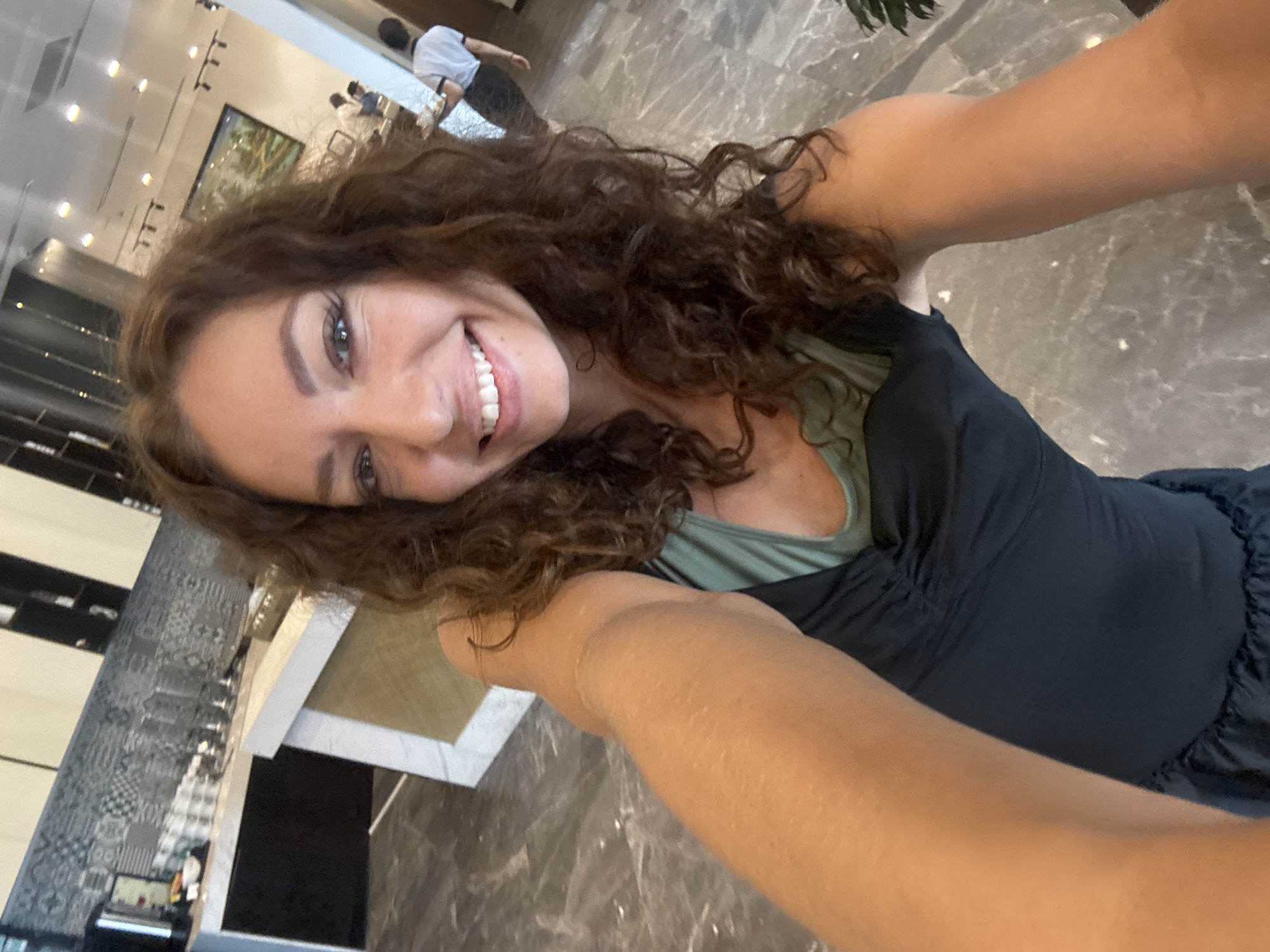 A woman with long, curly brown hair is taking a selfie indoors. She is smiling and wearing a dark-colored top with a visible green undershirt. The background shows a brightly lit, modern interior with marble floors. Other people are visible in the distance, one wearing a white shirt and dark pants. There are counters or tables in the background, along with light fixtures and other architectural details. The location appears to be a commercial space, possibly a hotel or mall in Đà Nẵng, Vietnam.