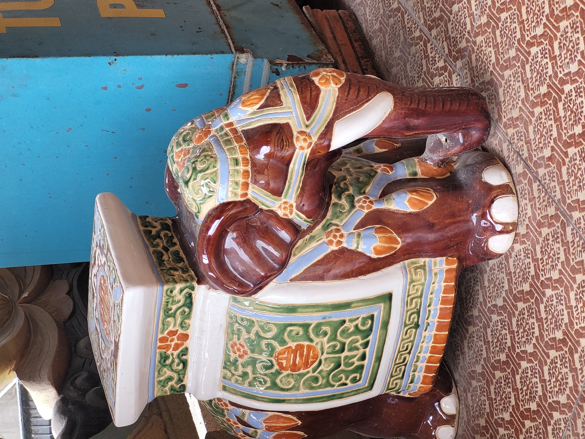 A ceramic elephant stool with intricate colorful patterns is displayed in an outdoor setting in Da Nang, Vietnam. The stool is predominantly brown with white accents on its tusks and feet, and features decorative panels on its back and head adorned with green, orange, and blue floral and geometric designs. The elephant's body is draped with ornate fabric-like coverings. The stool sits on a patterned tile floor with reddish-brown geometric motifs. In the background, a tall, weathered blue structure with yellow lettering is visible, suggesting it might be part of a shop or outdoor market stall. The overall scene is bathed in natural light.