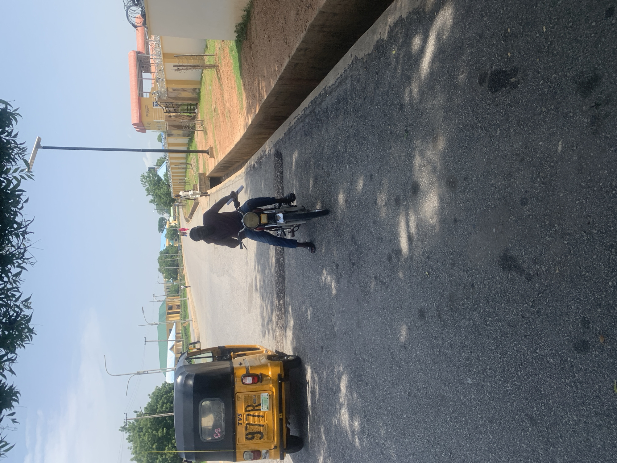 This is a photograph of a person riding a motorcycle on a paved road. The person is wearing a brown shirt and blue jeans. The motorcycle is dark colored. To the left of the motorcycle, a yellow and black three-wheeled vehicle, likely an auto-rickshaw, is parked. In the background, there are buildings with red and yellow facades, trees, and streetlights. The sky is clear and blue, suggesting a sunny day. The scene appears to be an outdoor urban or semi-urban environment. The photograph is taken from a slightly elevated angle and is oriented vertically. The shadows cast by the trees indicate it is likely daytime. The license plate of the auto-rickshaw is partially visible and appears to read "977R".